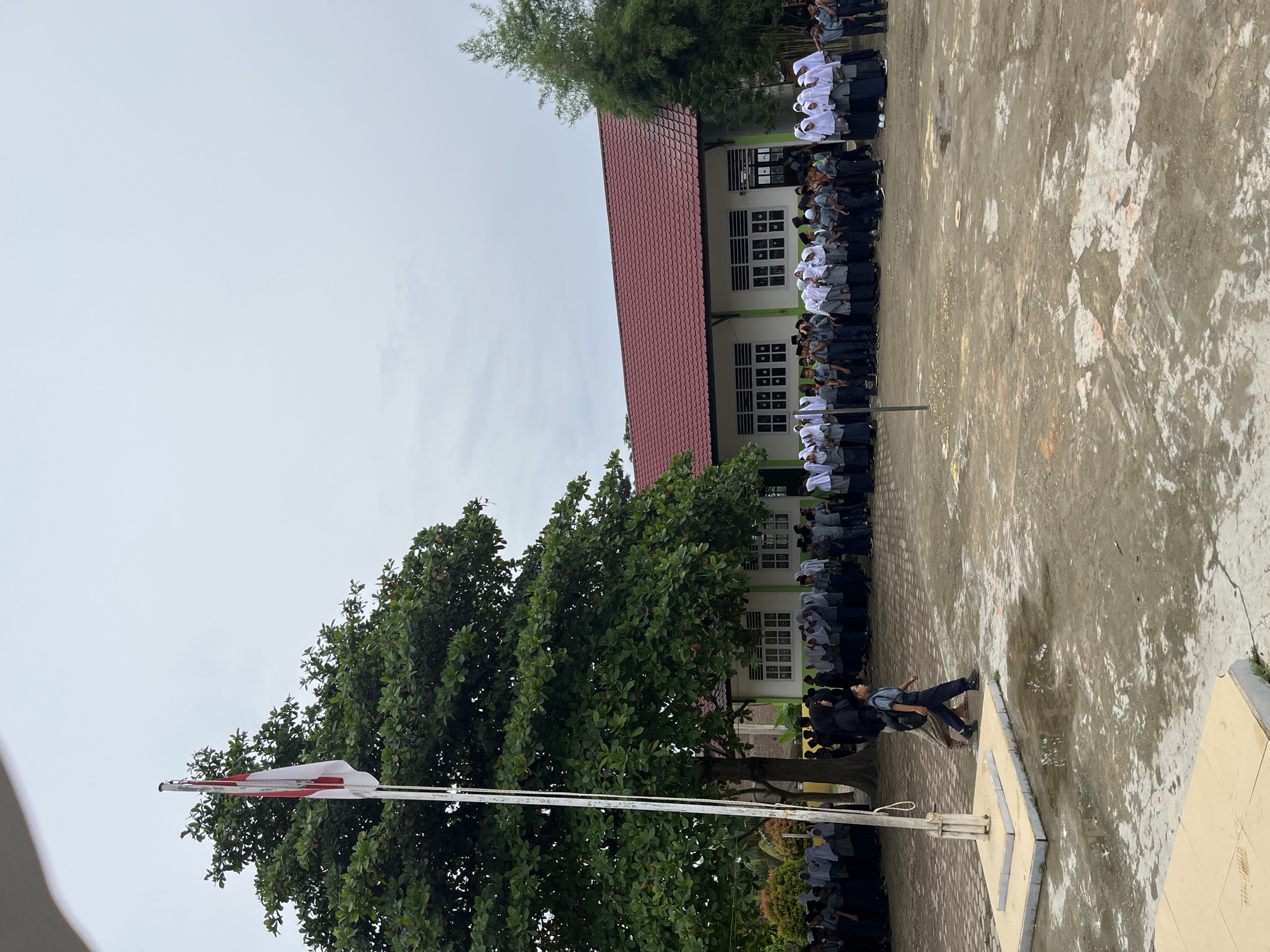 In Langnibong, Indonesia, a school gathering is underway on an overcast day. A group of students, predominantly dressed in gray uniforms and white headscarves, stand in formation near a light green building with a red tiled roof. Some students are in a line, while others are dispersed. A flag pole with the Indonesian flag, red and white, stands prominently to the left, partially obscured by a large, leafy tree. Two boys are walking across the wet, muddy courtyard in the foreground, one carrying a backpack. The environment is a schoolyard with puddles on the ground, indicating recent rain.