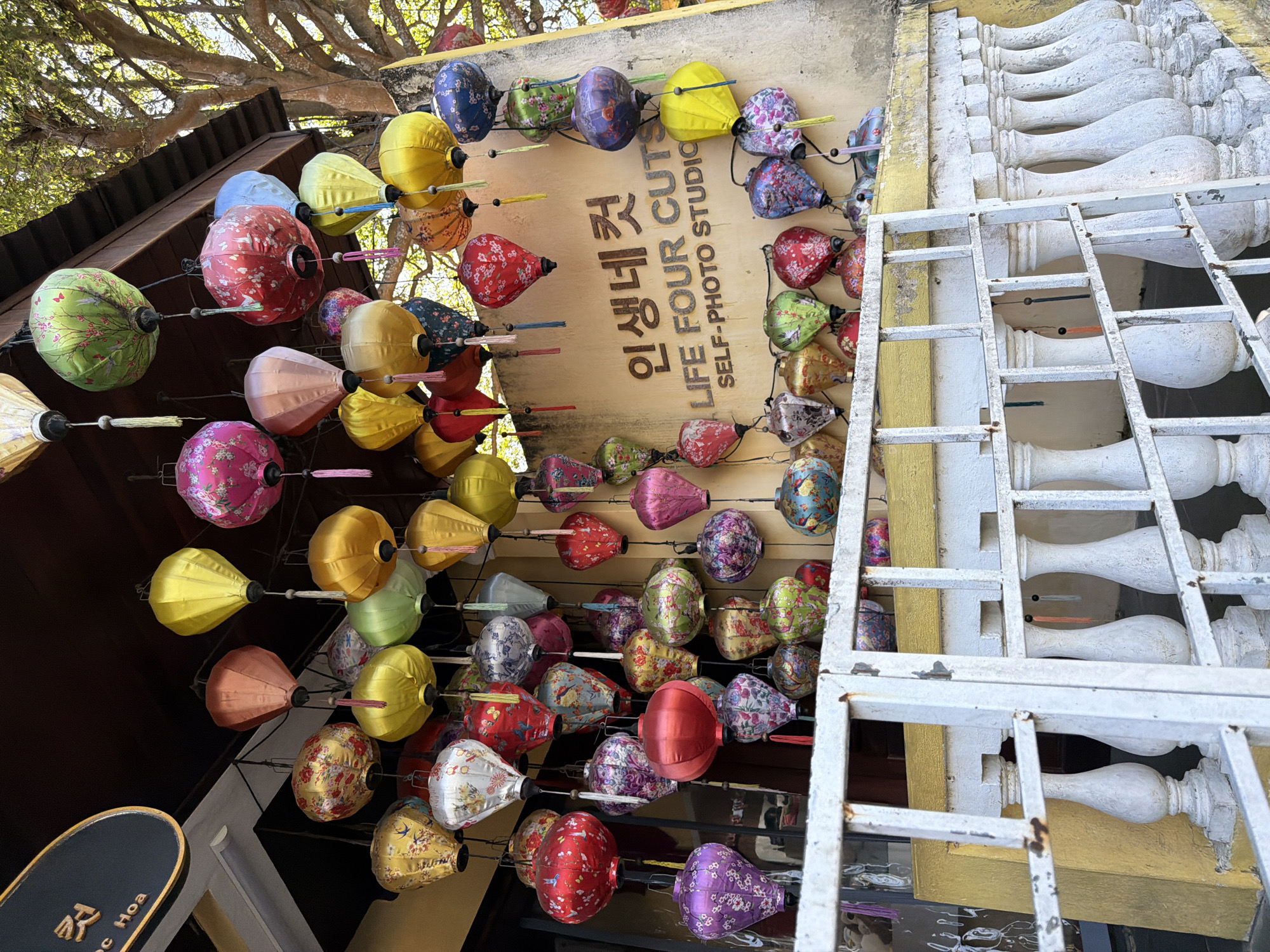 The image shows a storefront in Hoi An, Vietnam, decorated with numerous colorful silk lanterns. The lanterns are of various shapes and sizes, with intricate floral patterns and tassels. They are hung from the eaves of a building, creating a vibrant and festive display. The building itself appears to be a self-photo studio, as indicated by the signage in Korean and English that reads "인생네컷 LIFE FOUR CUTS SELF-PHOTO STUDIO." The studio is situated along a street lined with a white, ornate railing. In the background, lush green trees with dappled sunlight filtering through their leaves suggest a bright, sunny day. The overall scene is one of traditional Vietnamese aesthetics combined with a modern business.
