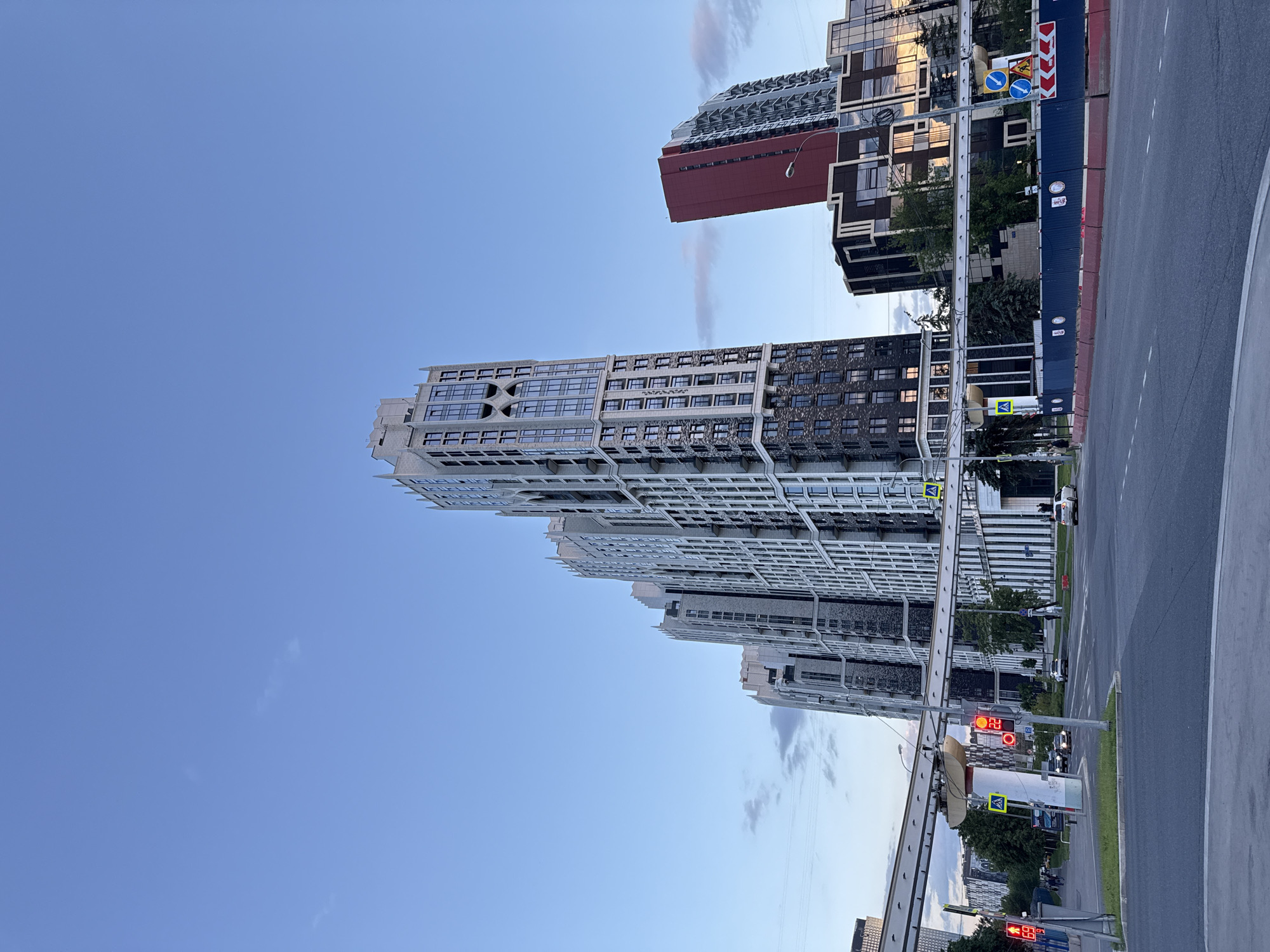 This image captures a modern urban street scene in Moscow, Russia, under a clear, bright blue sky with a few wispy clouds, suggesting late afternoon or early evening.
The dominant features are several tall, contemporary high-rise buildings lining a street. On the left, a prominent light-colored building with an intricate facade, numerous windows, and architectural projections stands out. To its right and slightly behind, another building features a darker, textured surface, possibly in shades of grey and black. Further down the street, a building with a distinctive red section is visible, contrasting with the other structures.
In the foreground on the right, a multi-lane asphalt road curves gently, with several cars stopped. A tall blue construction barrier runs alongside the road, separating it from the buildings and obscuring the immediate ground level. Several red traffic lights are active; one in the lower right corner displays "65" (seconds remaining), and another further up the road shows "29". Visible road signs include a blue circular sign indicating a turn or straight movement, a red and white warning sign (possibly for a barrier or hazard), and a yellow diamond-shaped pedestrian crossing sign. A few pedestrians are subtly visible near the crossing.
The scene depicts a typical urban environment, characterized by modern architecture, active traffic, and clear weather. The overall impression is one of a developed, busy city district.