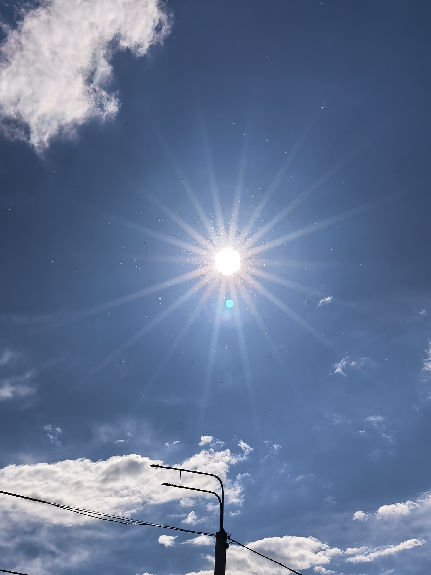 The image depicts a bright, sunny sky over Sterlitamak, Russia. The sun, centered in the upper middle, emits an intense starburst effect with prominent rays and a noticeable teal lens flare directly below it. Numerous small, bright specks, possibly dust or pollen illuminated by the sunlight, are scattered across the sky. White cumulus clouds are visible: a large, dense formation occupies the upper left, while smaller, scattered clouds appear in the mid-left and lower right. A long, horizontal band of clouds stretches across the lower portion of the sky. In the lower center, a dark silhouette of a street lamp pole and its two curved arms is present, along with several dark power lines extending horizontally from the pole. No people are visible.