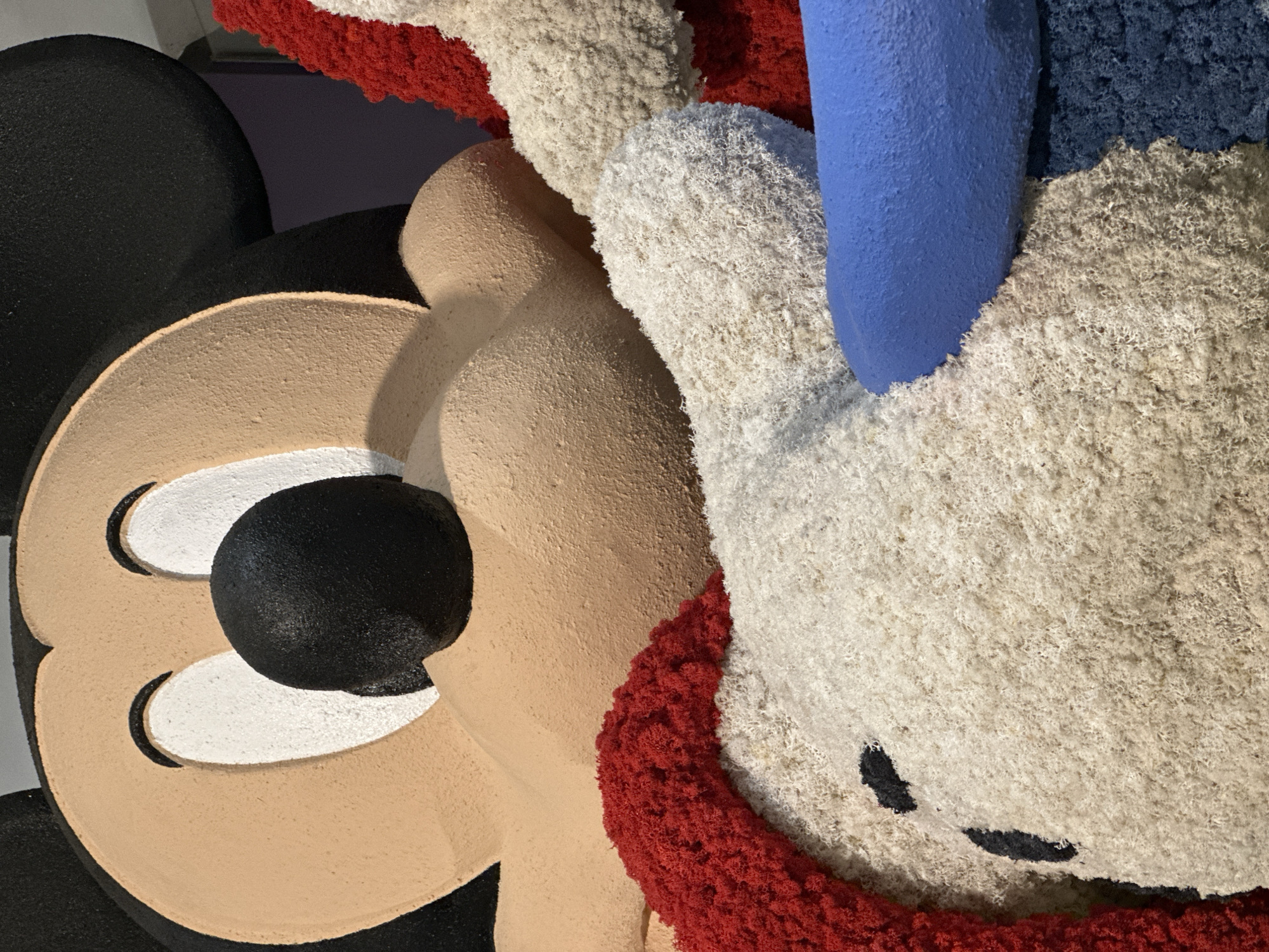 This is a close-up photograph of a large, textured sculpture of Mickey Mouse's face and upper body. The sculpture has a rough, granular texture, and its colors are muted. Mickey's iconic features are prominent: his large, circular ears are black, his face is a tan color, and his eyes are white with black pupils. His nose is a large, black sphere. He appears to be wearing a red and white striped outfit. The sculpture is part of an outdoor display, and the background is out of focus, showing some indistinct shapes and colors that suggest a park or public space. The image was taken at the Singapore Zoo.