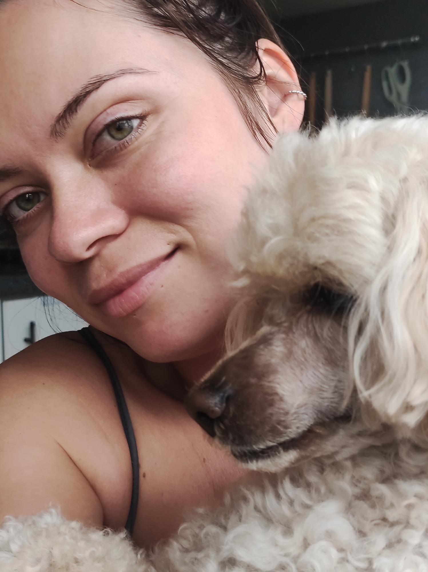 A close-up selfie shows a woman with light skin and green eyes, her left side facing the camera. She has dark hair pulled back and wears a thin strap top. A light-colored poodle is nestled against her right shoulder, with its head resting on her chest. The background is dark and out of focus, with some objects that appear to be scissors on a rack. The image was taken indoors. There is no discernible location in the city of Chaikovsky, Russia.