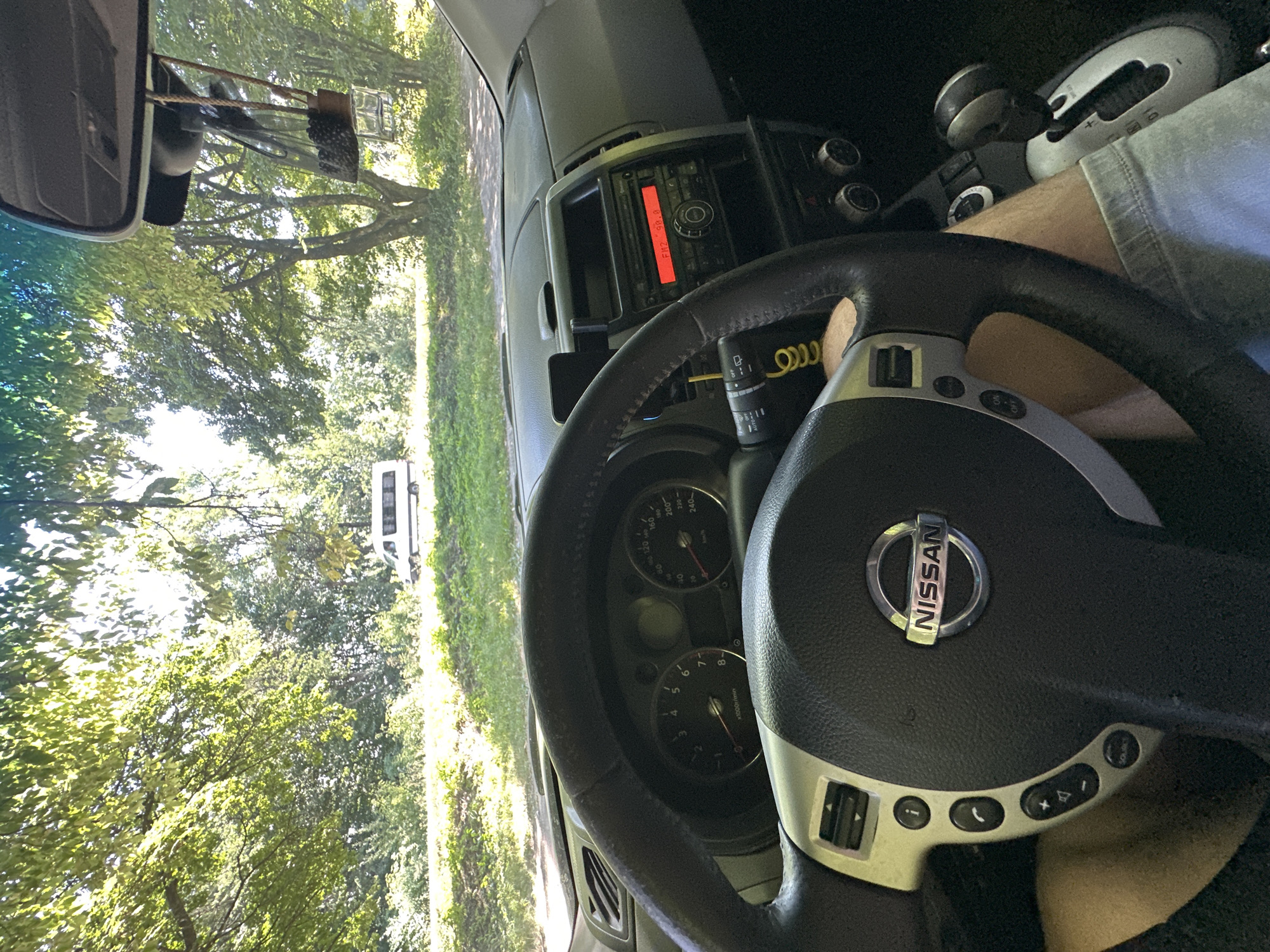 The image provides an interior view from a Nissan vehicle, looking forward through the windshield. A person's legs, clad in light-wash denim shorts, are visible in the lower right and left portions of the frame, indicating they are the driver. The steering wheel, displaying the Nissan logo, is prominent in the foreground, with dashboard controls and a radio with an illuminated red display showing "FM2 99.8" to the right. A white minivan is observed driving on a road in the distance, surrounded by lush green trees and foliage. The scene suggests the vehicle is parked or driving on a road bordered by a natural, wooded environment. The location is identified as Shatava, Ukraine.