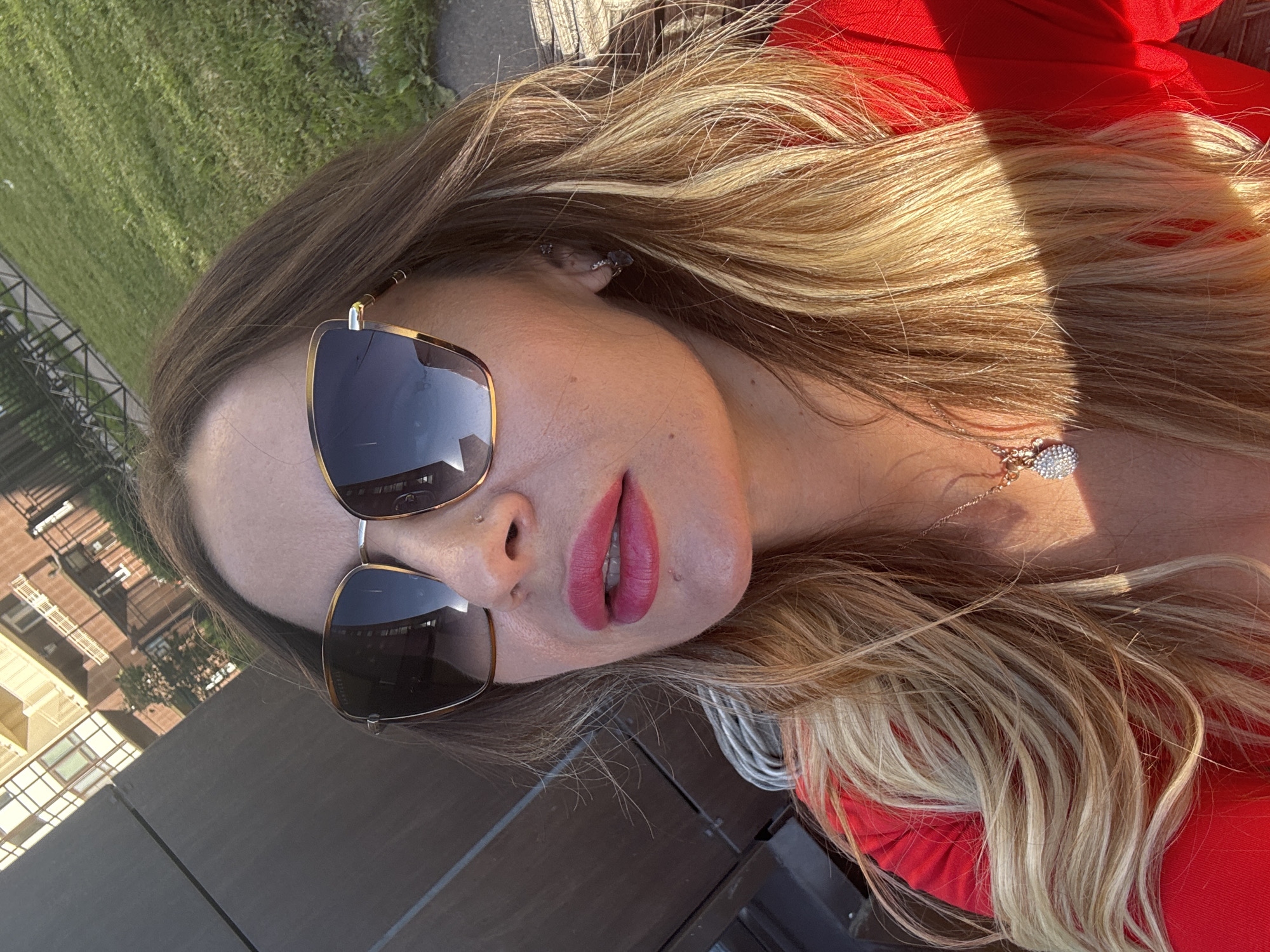 A woman is taking a selfie outdoors, with her head and shoulders filling the frame. She is wearing large, tortoiseshell sunglasses and bright pink lipstick. Her blonde hair is styled in loose waves and falls over her shoulders. She is also wearing a bright red top and a delicate necklace with a sparkling pendant. In the background, a building with a modern architectural style and balconies can be seen, along with a patch of green grass and a fence. The lighting suggests it is a sunny day. The presence of the building with balconies is characteristic of residential architecture found in Saint Petersburg, Russia.