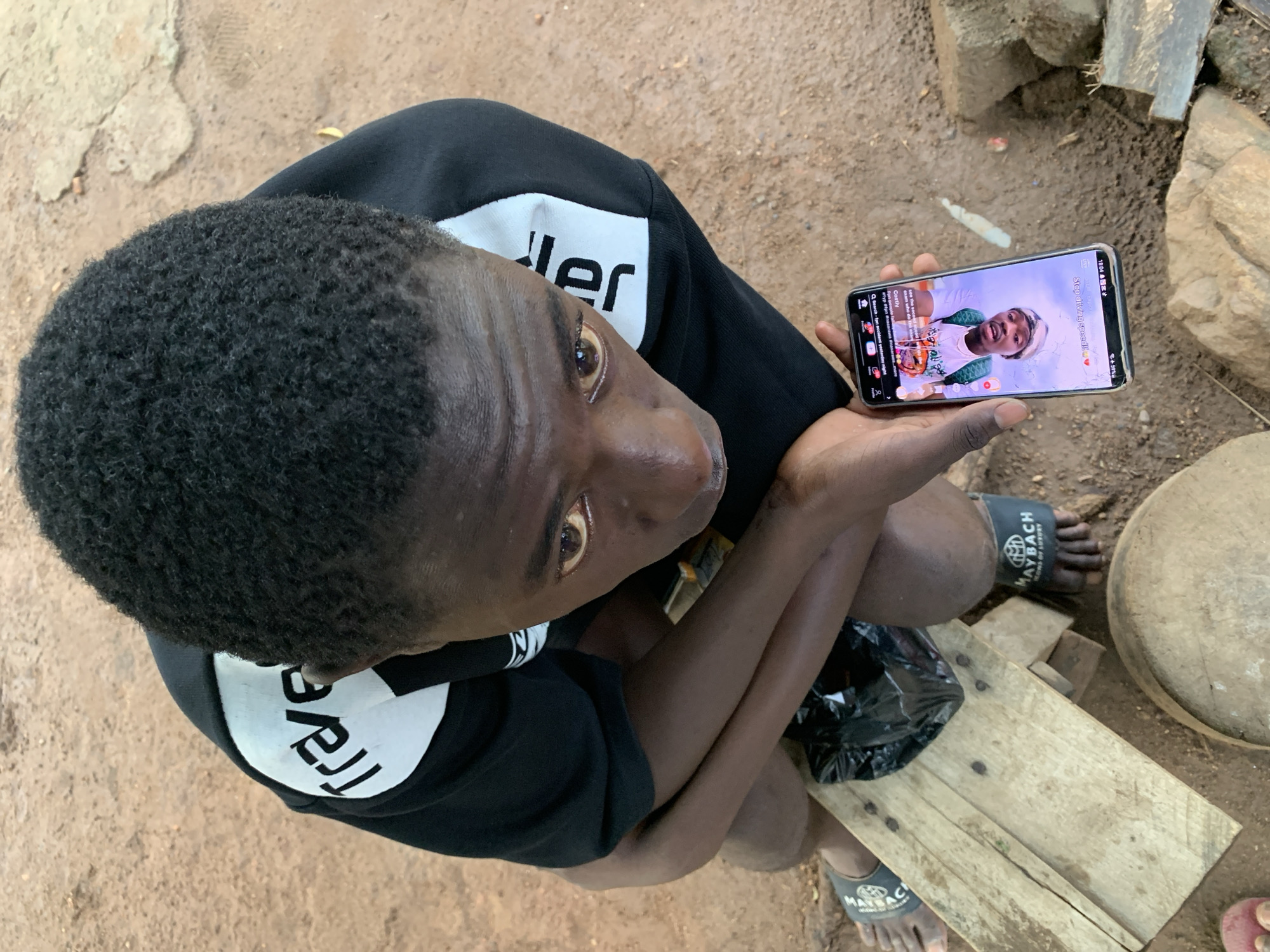 A top-down, high-angle shot shows a dark-skinned person, likely male, with short, dark, natural hair, closely cropped to the head. The person is wearing a black hooded sweatshirt with white lettering on the front. The person is seated, with knees drawn up and arms crossed in front of them, holding a smartphone in their right hand. The phone displays a video or live stream, with a person visible on the screen. The phone screen shows text including "Stop driving speed!!", "Costly," and a search query. The person is wearing black sandals with white logos.
The environment consists of dirt ground and stone features. There is a wooden plank and a round wooden object, possibly a stool or drum, to the lower right of the person. The image appears to be taken outdoors in a rural or less developed area, possibly in the city of Akwanga, Nigeria.