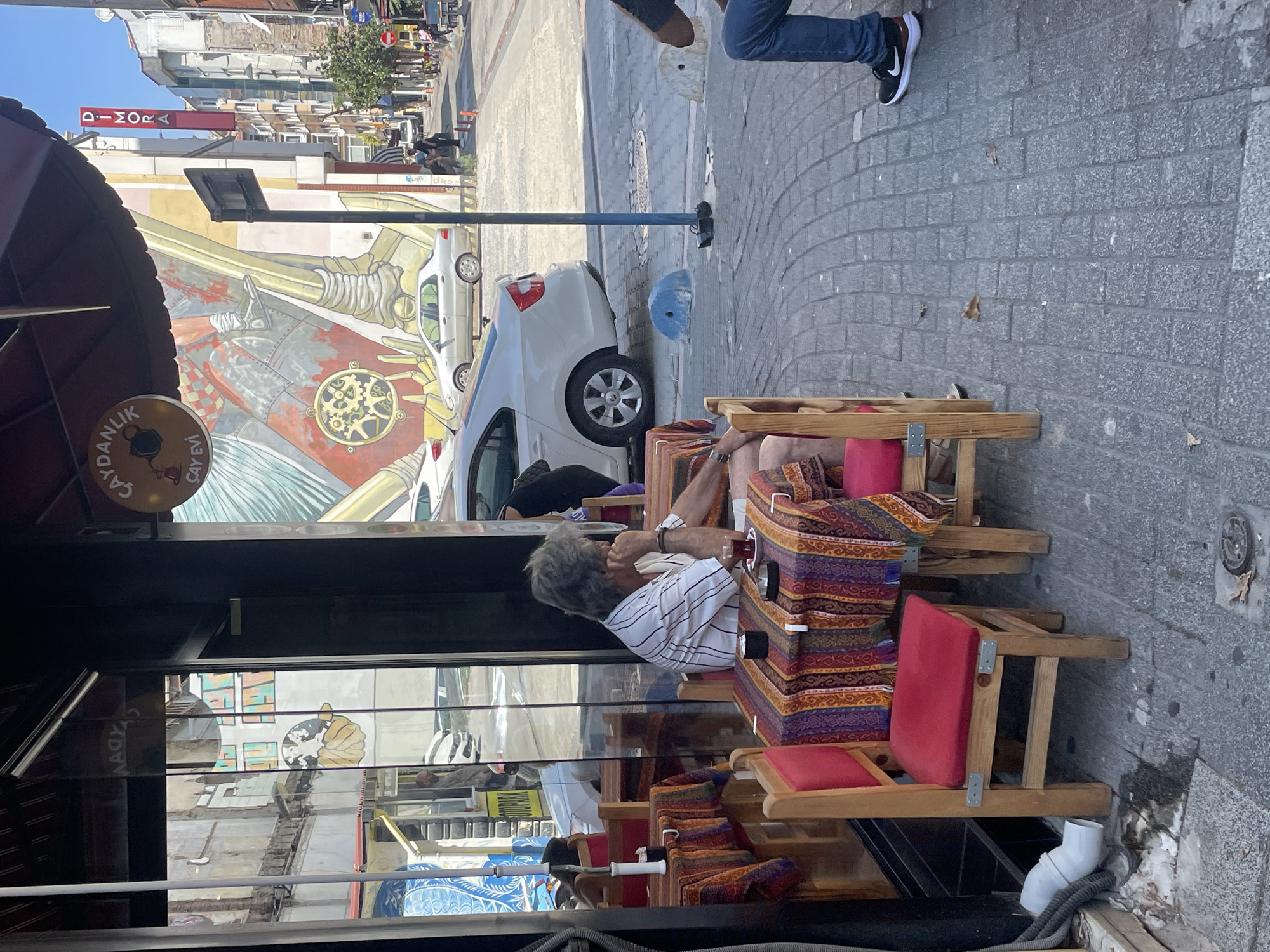 The image depicts an outdoor seating area of a cafe in Kadıköy, Turkey. A man with graying hair, wearing a striped shirt, is seated at a small table covered with a vibrantly patterned tablecloth. He is holding his chin with his hand and appears to be in thought. A small cup of dark liquid, likely tea, and an ashtray are on the table. The cafe's interior is visible through the glass, showing reflections of buildings and a mural. Across the street, there are more buildings, parked cars, and people walking. The ground is paved with grey cobblestones. A red sign with white lettering saying "DİMORA" is visible on a building in the background.