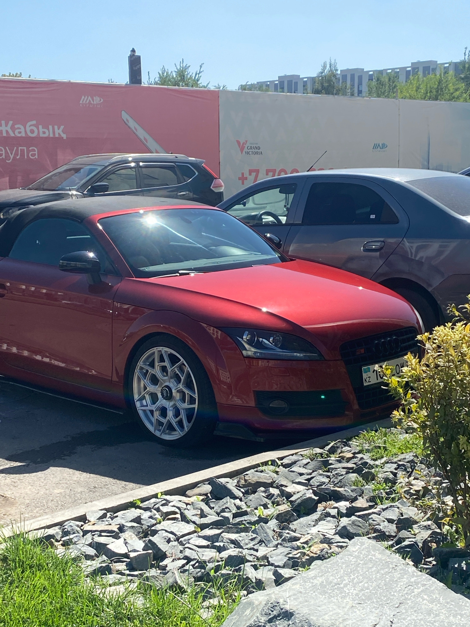The image shows a red convertible Audi TT parked in what appears to be a construction or development area in Astana, Kazakhstan. In the background, a large pink banner displays text in Cyrillic, possibly advertising a development called "GRAND VICTORIA" with a phone number starting with "+7 700". Another banner behind it is light-colored with a logo that includes the letters "HAD". Several modern buildings with white facades and dark windows are visible in the distance, suggesting an urban environment. The red Audi is in the foreground, with its convertible top down. Behind it, a dark-colored SUV is partially visible, and further back, a dark sedan is parked. The ground is paved with asphalt and bordered by rocks and some green foliage. The sky is clear and blue. There are no people visible in the image.