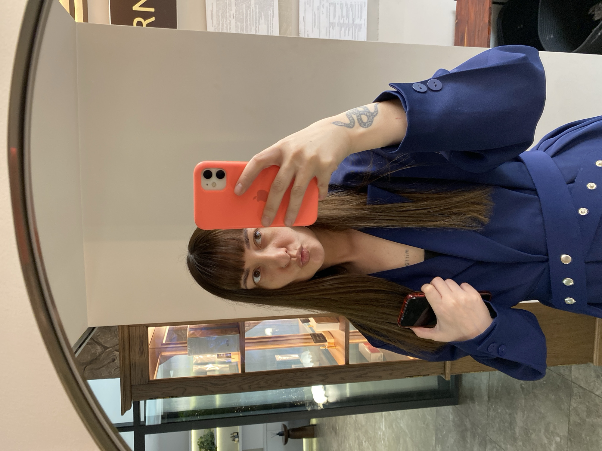 A woman is taking a selfie in a mirror. She is wearing a blue blazer-style top. She is holding a phone with a bright orange case up to the mirror with her left hand, and another phone in her right hand. The mirror is oval and framed. Behind her, in the reflection, is a display case with shelves containing various objects. The background suggests a store or commercial space, potentially in Votkinsk, Russia.