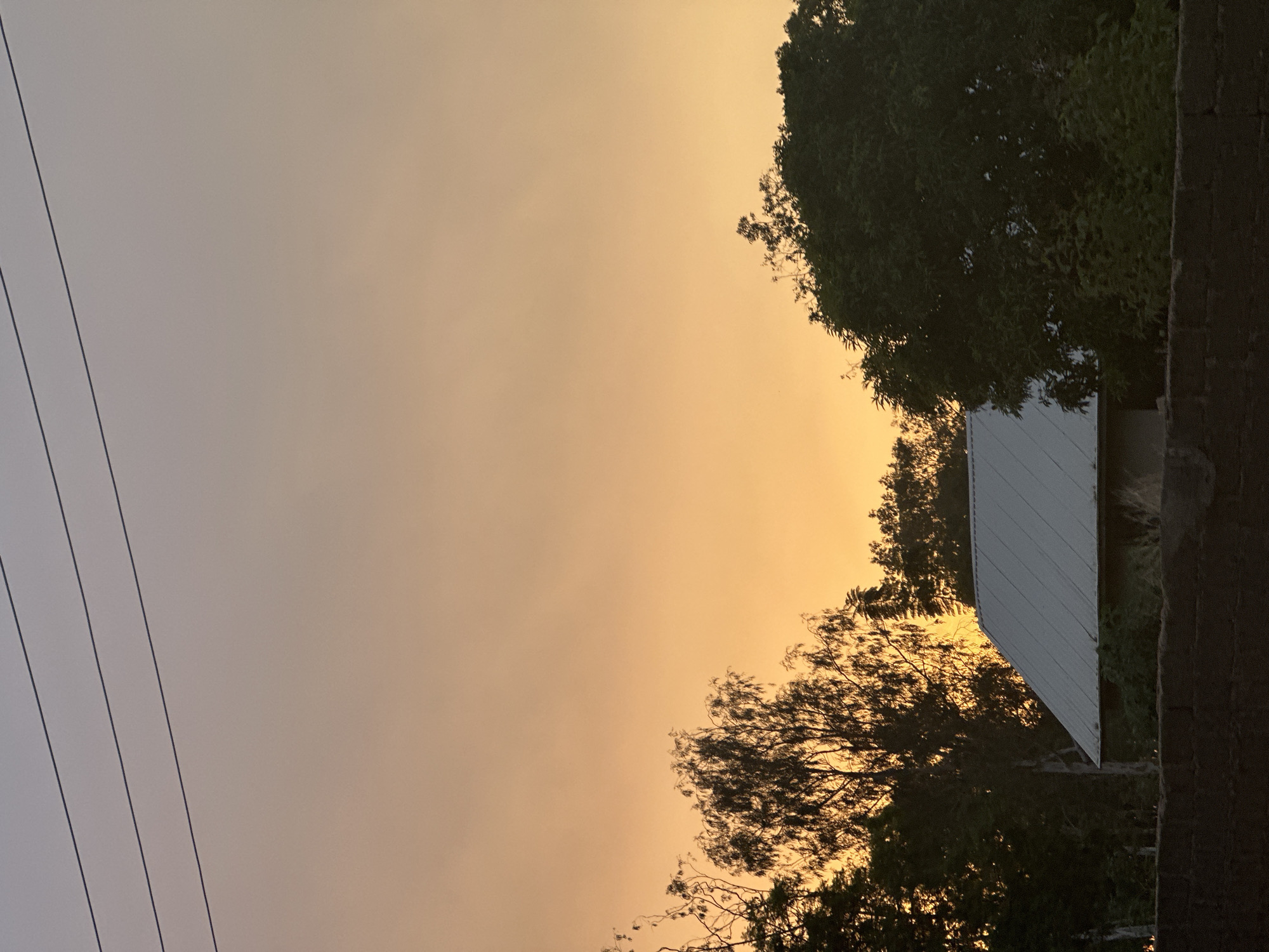 A dusk view taken in Bukuru, Nigeria, showcases a hazy orange and yellow sky filling most of the frame. Power lines run vertically along the left edge. Trees with dense foliage are silhouetted against the sky on the right side. A white corrugated metal structure is seen partially obscured by the trees, near a dark brick wall.