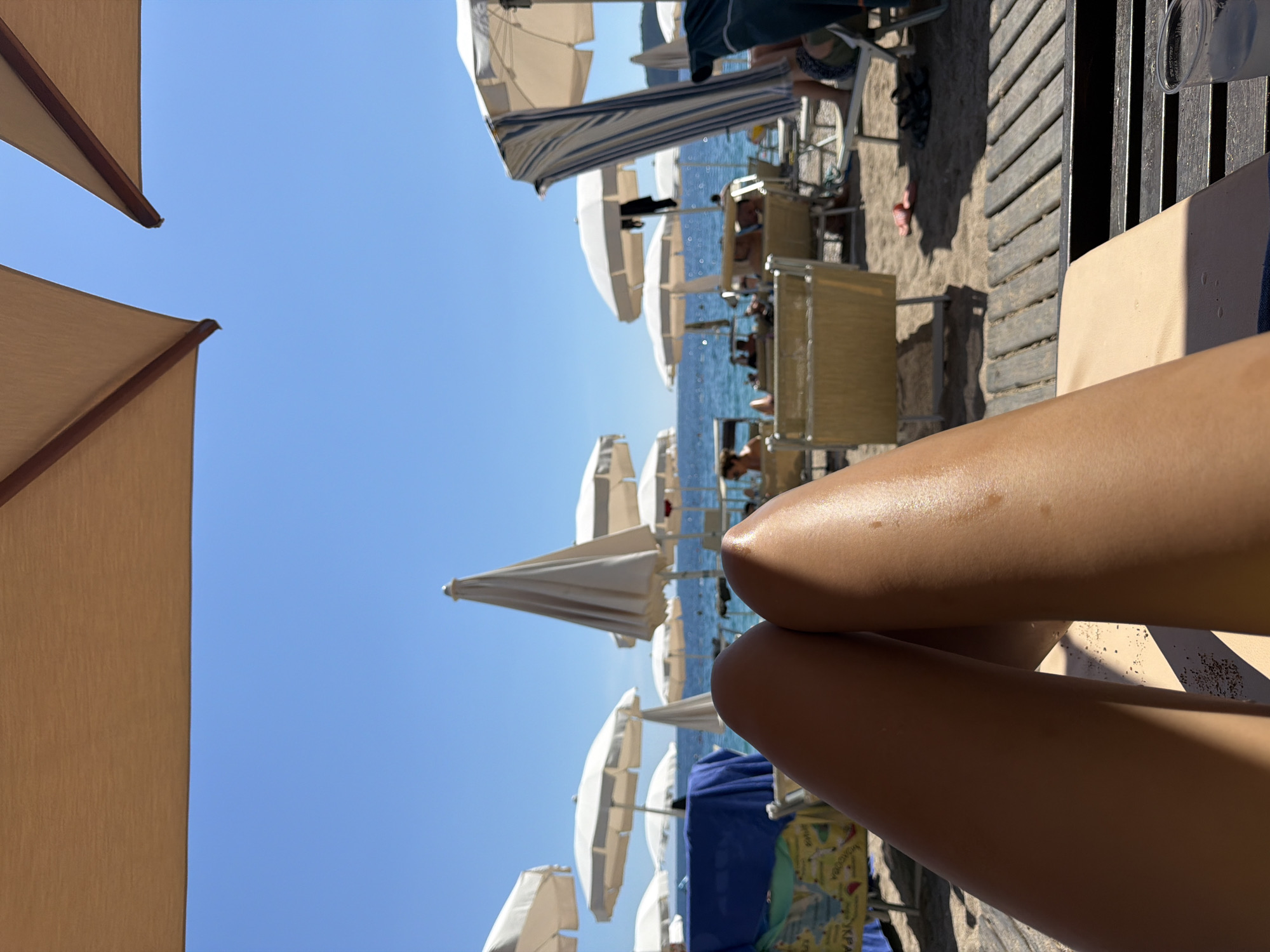 The image depicts a first-person perspective of someone relaxing on a beach. The viewer's legs are prominently in the foreground, positioned centrally and slightly to the left. The legs are tanned and appear to be resting on a light-colored surface, possibly a beach chair or lounger.
In the background, a beach scene unfolds under a clear blue sky. Several white beach umbrellas are visible, providing shade. Beyond the umbrellas, the ocean stretches to the horizon, with calm water. Beach chairs and loungers are scattered across the sandy area, some occupied by people.
To the left, a blue towel or blanket is draped over a beach chair. The ground consists of sand and wooden planks. The overall atmosphere suggests a sunny day at a beach resort. The location is not specified within the image itself.
