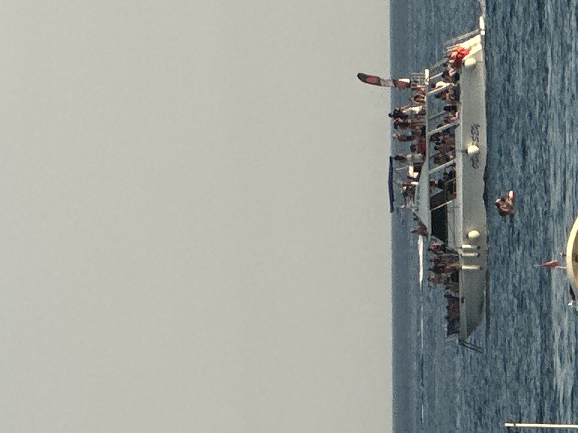 A white catamaran with "ODYSSEY" written on its side is sailing on a blue sea. There are many people on the boat, some standing on the upper deck and some sitting inside the cabin. A red and black flag is attached to a pole on the back of the boat. The sky is clear and pale. In the foreground, a person is swimming in the sea with a red float. The scene takes place on the coast of Boreti, Montenegro, as indicated by the context.