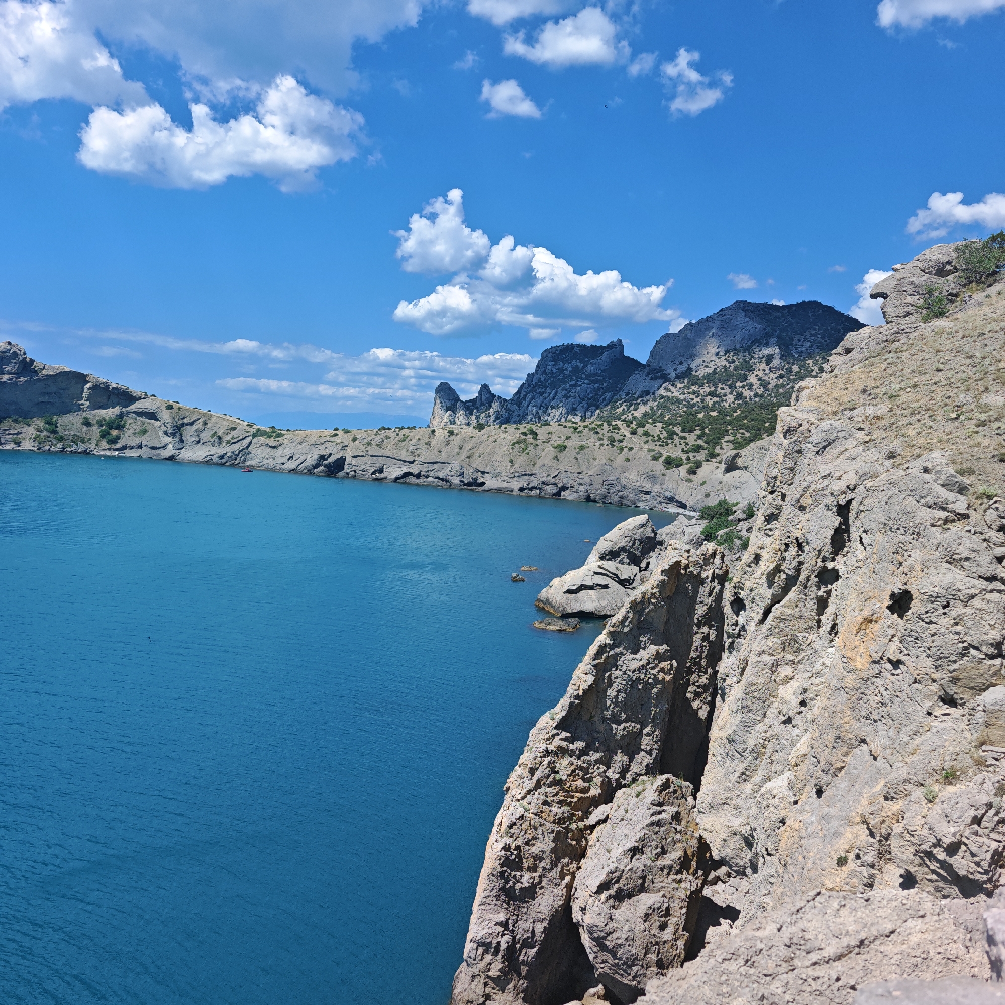 The image shows a rugged coastline with steep, rocky cliffs overlooking a vibrant blue sea under a bright, cloud-dotted sky. In the distance, a series of jagged mountain peaks rise from the land. The foreground is dominated by a rocky, sun-drenched cliff face. The sea is calm with gentle ripples. There are no visible people in the frame. The scene is likely a natural landscape, possibly a hiking trail or viewpoint in a coastal area. The location is identified as the city of Sudak, Ukraine, which is known for its dramatic coastline and mountainous terrain.
