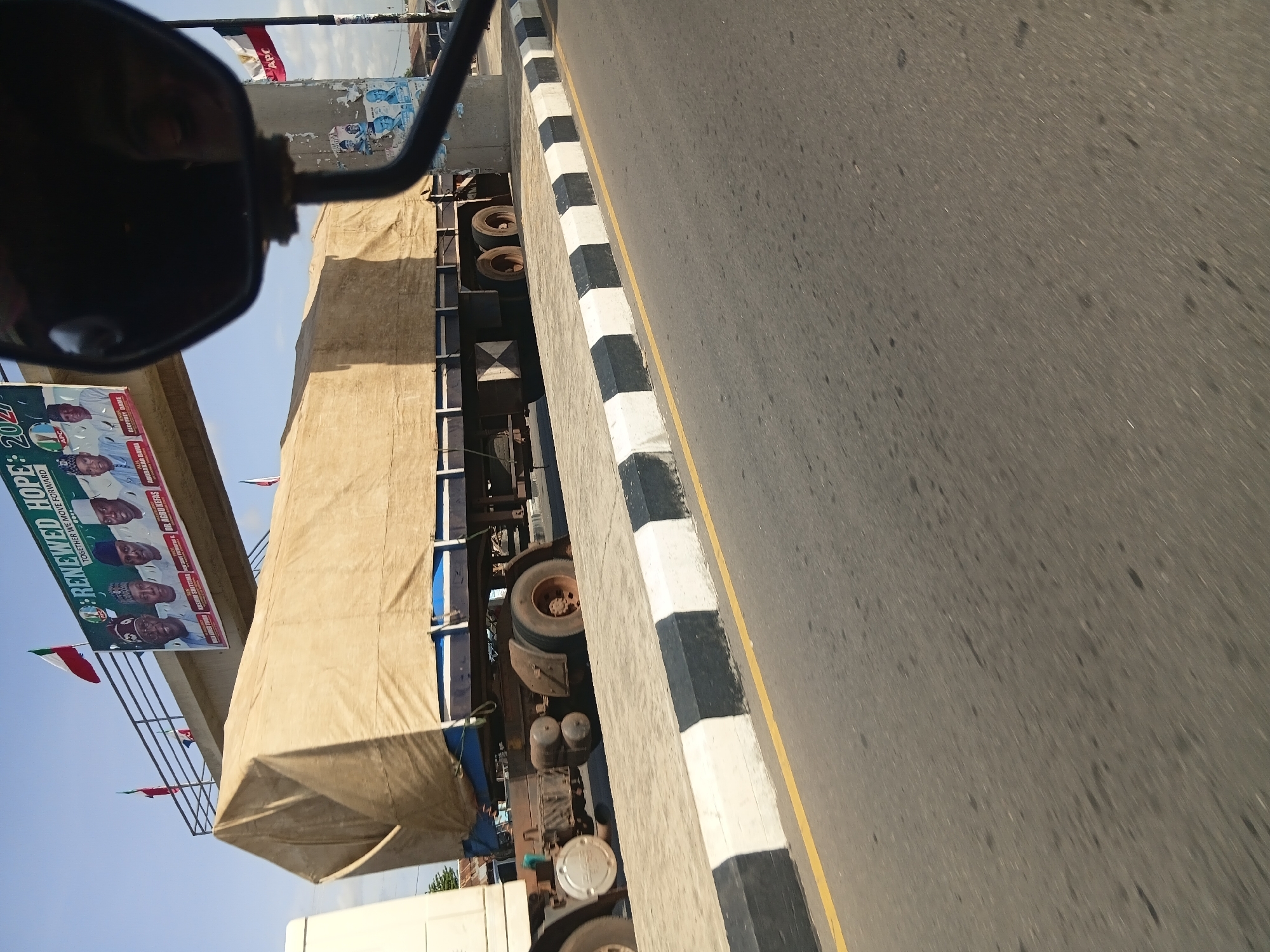 A medium shot captures a cargo truck partially covered with a beige tarp, driving on a road in Jalingo, Nigeria. The truck has visible wheels and a blue trailer. The scene is set against a clear blue sky with a large banner overhead featuring portraits of several men and the words "RENEWED HOPE 2027 TOGETHER WE MOVE FORWARD". Several flags are visible, including Nigerian flags and flags of a political party. A concrete barrier with alternating black and white sections separates the road from a sidewalk. A reflection of a person's face is visible in a rearview mirror in the foreground. The environment suggests an urban setting with a clear sky and roadside infrastructure.