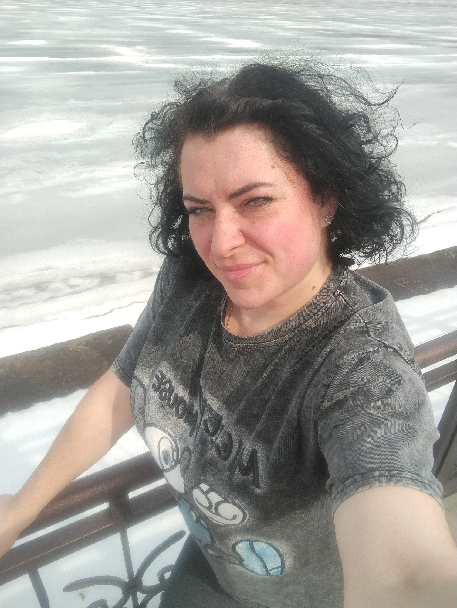 A medium shot depicts a woman with curly, dark hair, a grey t-shirt with a cartoon design, and reddened cheeks, seemingly from the cold. She is positioned at an elevated viewpoint, possibly a bridge, with a railing visible in the foreground. Beyond her, a partially frozen body of water stretches into the distance. Based on the appearance of the setting and the context, this is likely in the city of Votkinsk, Russia.