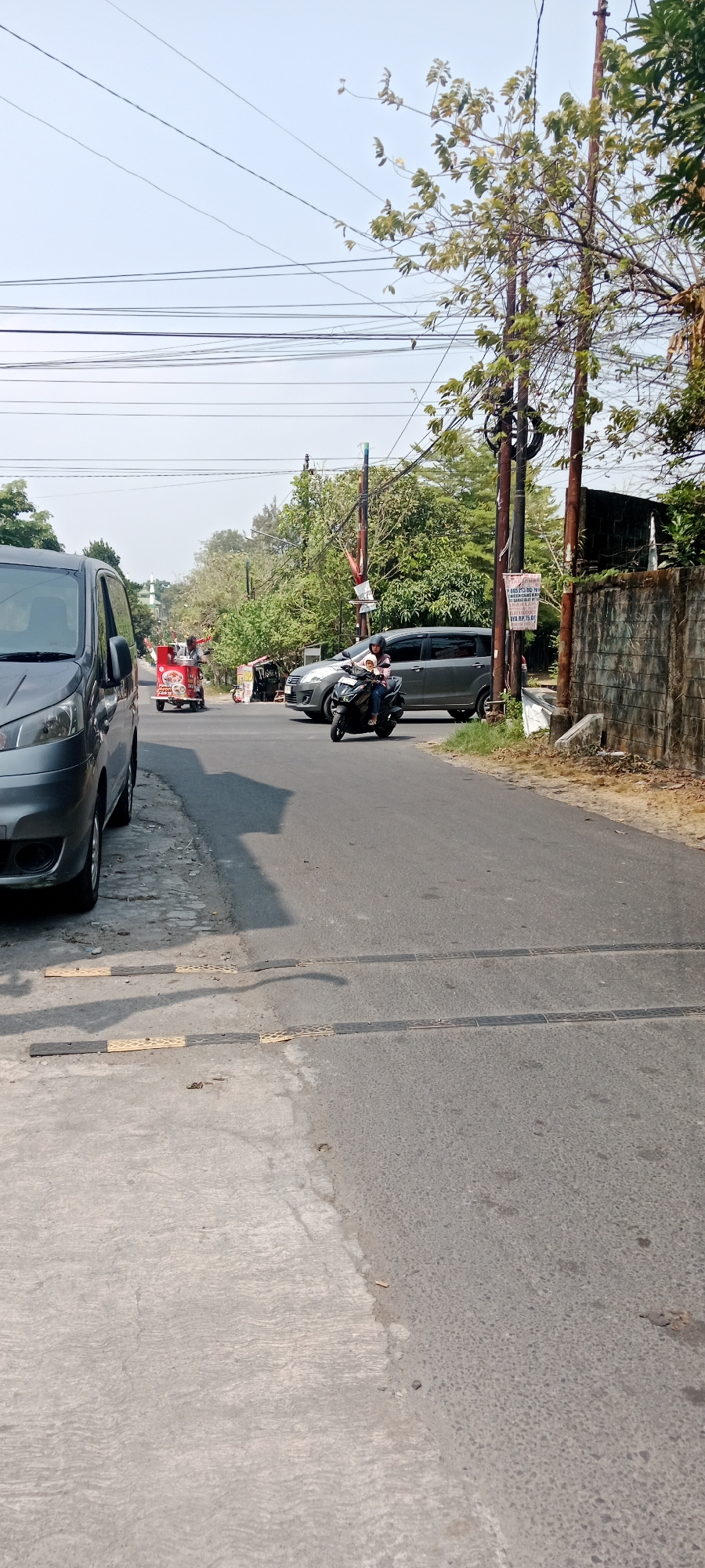 The image shows a street scene in Maguwoharjo, Indonesia, on a bright, sunny day. A grey van is parked on the left side of the road, partially obstructing the view of the sidewalk. A person on a black scooter, with a passenger, is riding on the right side of the asphalt road. Behind the scooter, a grey SUV is also on the road. Further down the road, a red food cart with a person standing next to it is visible. Overhead, numerous electrical wires are strung between poles. Trees with green foliage line the side of the road, and a textured brick wall is present on the right. The road has speed bumps. The sky is clear and light blue.