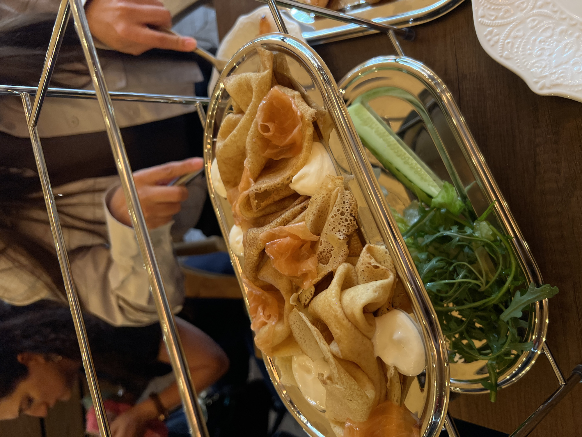 The image depicts two oval silver trays resting on a multi-tiered silver stand on a dark wooden table. The tray on the left contains rolled crepes filled with salmon and a dollop of white cream. The tray on the right holds green leafy vegetables and sliced cucumbers. Two individuals are visible, with one person wearing a light-colored shirt and using cutlery, and another person partially visible at the bottom left. The location may be in Yerevan, Armenia.