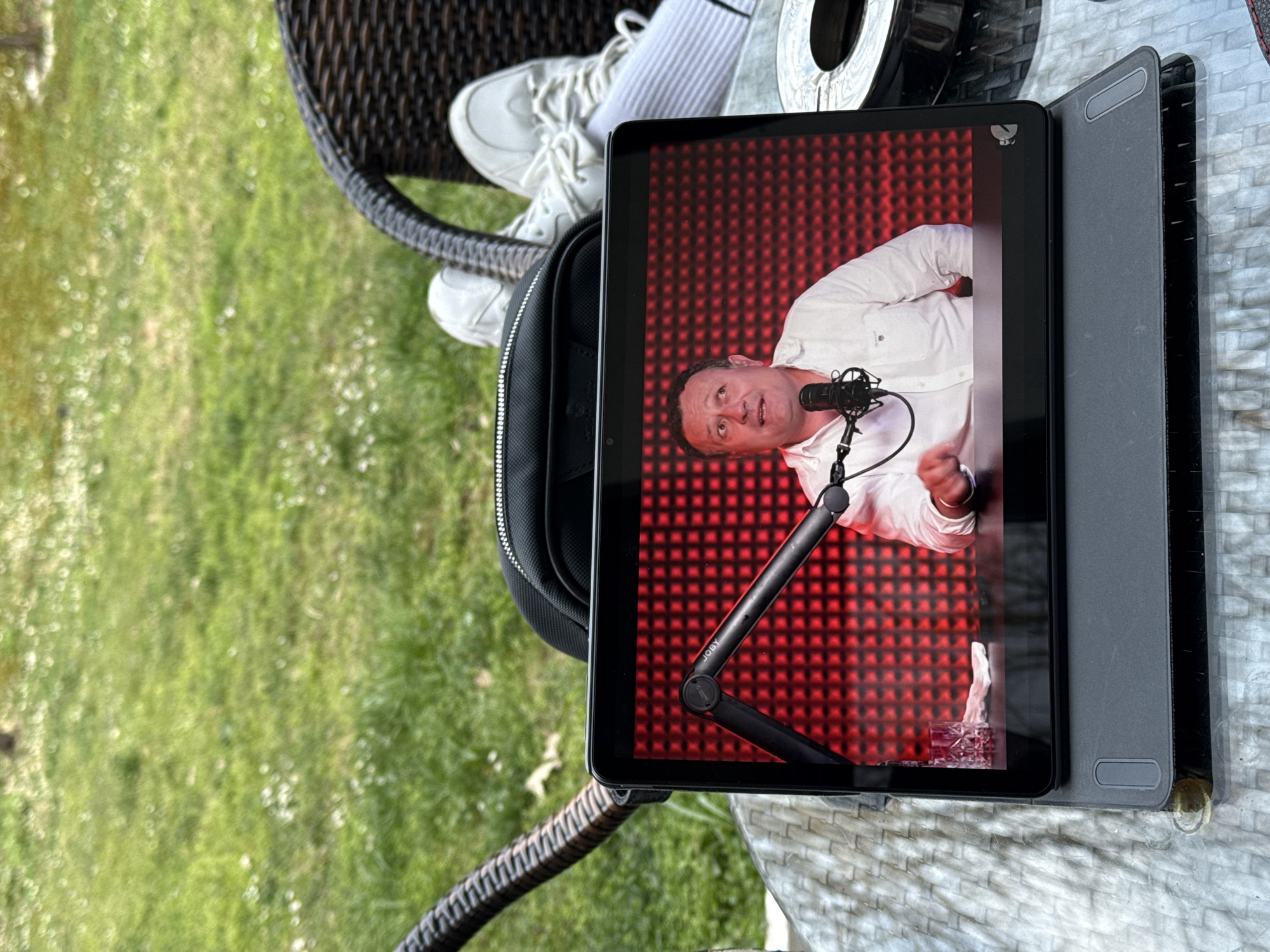 The image shows a tablet displaying a man speaking into a microphone against a red patterned background. The tablet is positioned on a table outdoors, with a wicker chair and green lawn visible in the background. Other objects nearby include a bag, shoes, and a silver metallic object. The location is outdoors, but it's not possible to discern if it is in Etropole, Bulgaria, based solely on the image.
