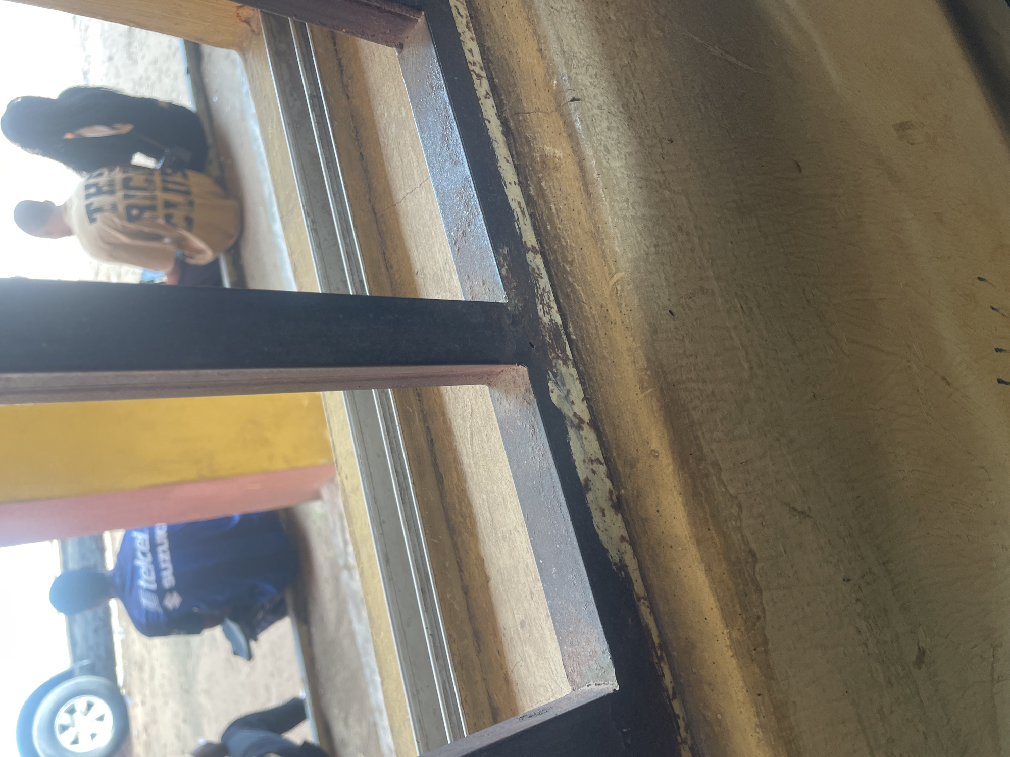 In Lokoja, Nigeria, a group of individuals are seen sitting outdoors. The scene, viewed through a metal grate, suggests a public space, possibly a waiting area or a roadside spot. In the foreground, a weathered, yellow painted surface with visible scuff marks and a metal grate dominates the lower portion of the image. Behind the grate, three people are visible. On the left, a person wearing a dark blue shirt with "telcel SUZUKI" logo is seated. To the right, two individuals are seated. One person wears a tan t-shirt with the words "THE RICH CLUB" printed on the back. Beside them, another person with long dark hair is also seated. In the background, a white vehicle's wheel and a portion of a yellow and pink wall are discernible. The overall environment appears to be an outdoor urban setting, likely during daylight.