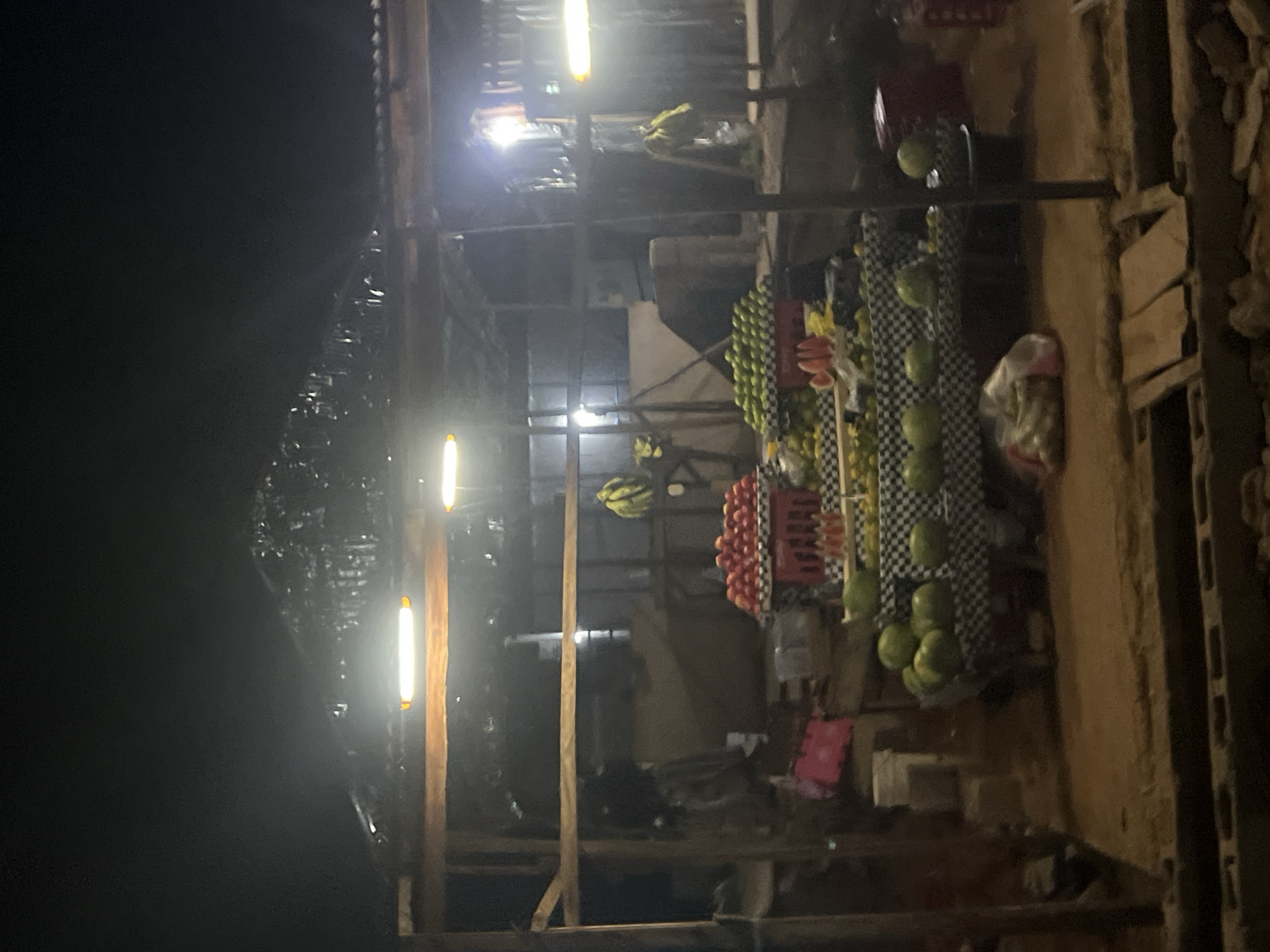 This image captures a dimly lit outdoor market stall at night, presumably in Durumi, Nigeria. The stall is constructed with rough wooden poles and a corrugated metal roof, illuminated by fluorescent lights. The primary focus is on a variety of fruits displayed for sale. Prominently featured are piles of red tomatoes in a red crate, green apples or limes, watermelons, and slices of watermelon. Bananas are also visible, hanging in bunches. The fruits are arranged on tables covered with checkered cloths. In the background, there are some bags and possibly crates, along with what appears to be another stall or structure behind the main display. The ground is sandy and uneven. The overall impression is of a bustling local market operating after dark.