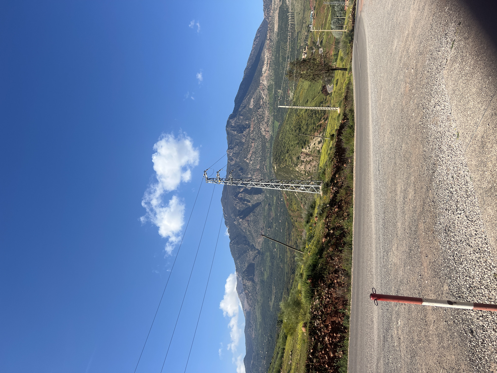 The image depicts a landscape in Ouaouizert, Morocco. A road runs along the right side of the frame, with a red and white barrier visible on the pavement. A mountain range is located on the left side of the road, with green vegetation covering its slopes. A utility pole and power lines are positioned in front of the mountain. The sky is bright blue with scattered white clouds.
