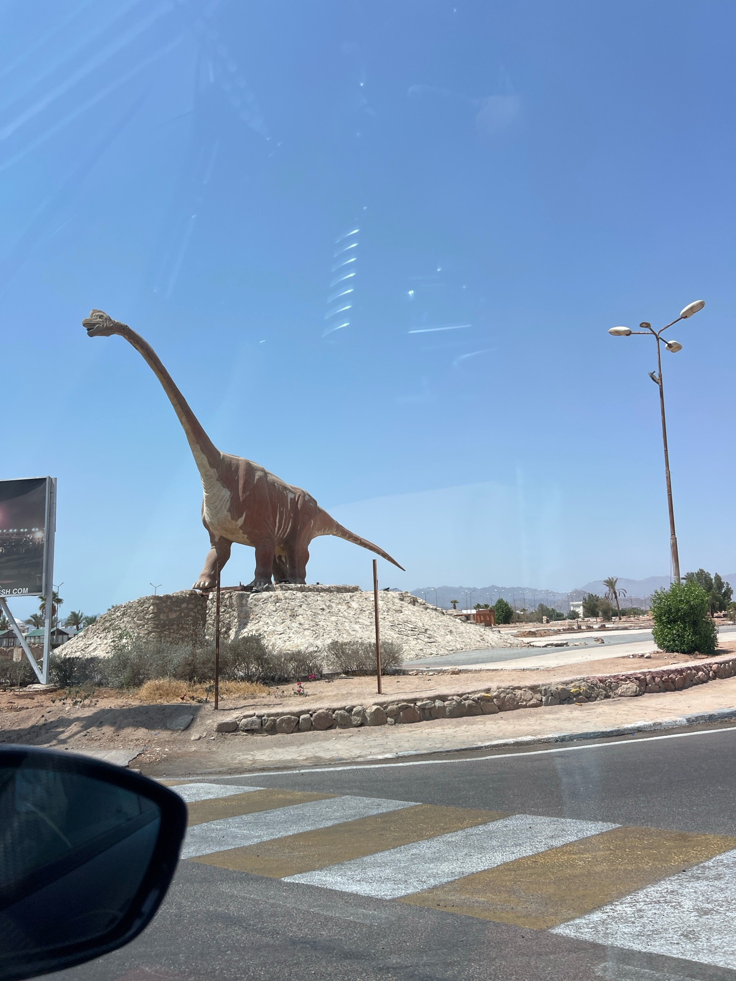 A large, brown statue of a Brontosaurus dinosaur stands on a rocky, artificial mound under a clear blue sky. The dinosaur is positioned to the left of the frame, facing right, with its long neck extended. To the right of the dinosaur, a multi-headed street lamp stands tall. In the foreground, a crosswalk with yellow and white stripes is visible on an asphalt road. A car's side mirror is reflected in the lower left corner of the image. In the background, there are trees, buildings, and mountains in the distance. This scene is located in Sharm El Sheikh, Egypt, specifically near the entrance to the city, marking a prominent landmark.