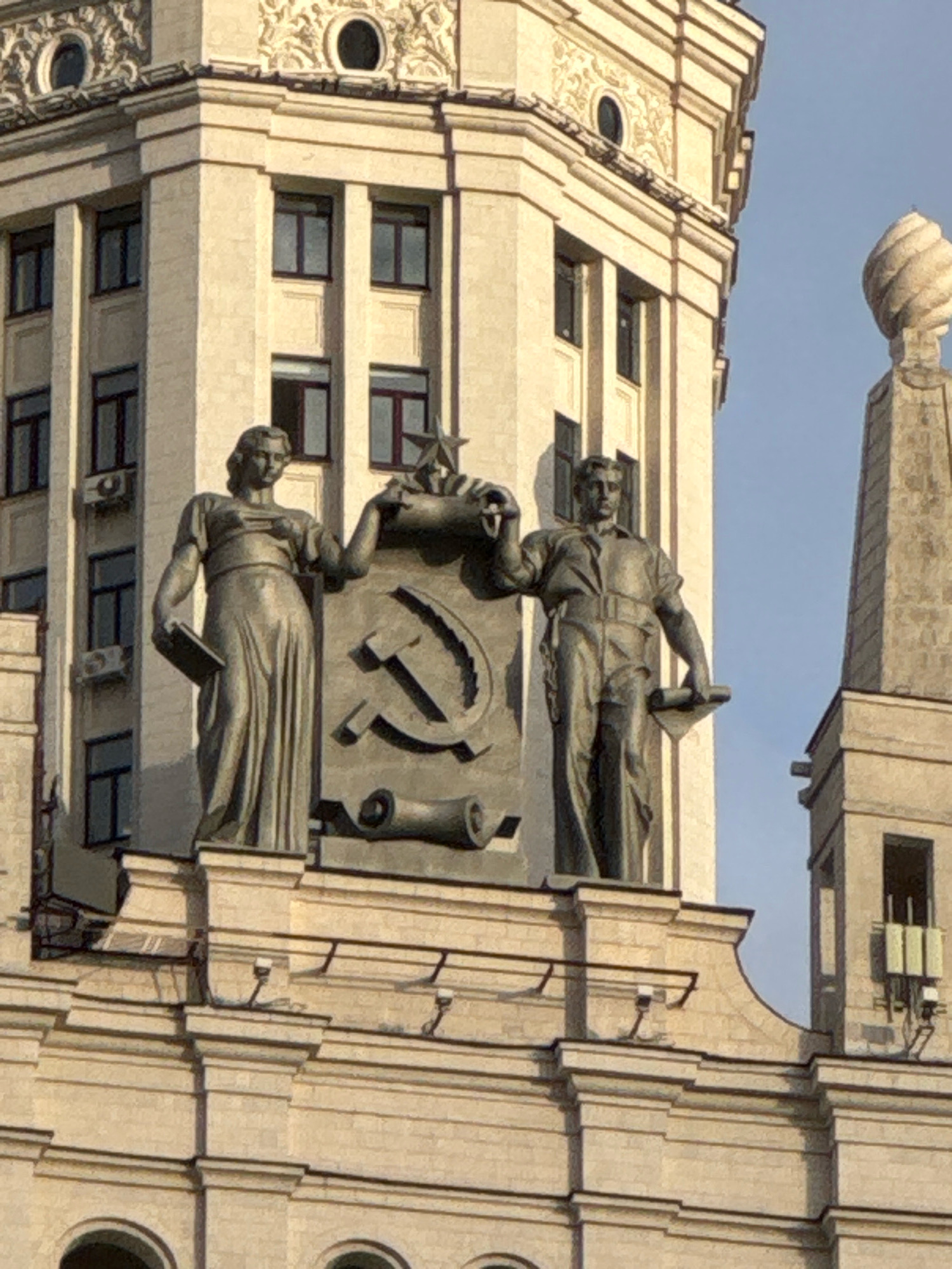 The image captures a close-up view of the Stalinist architecture of the Moscow State University (MSU) main building, a prominent landmark in Moscow, Russia. The foreground features two large, bronze statues atop the building's facade. The statue on the left depicts a woman in a flowing, classical-style robe, holding a book in her left hand and looking forward. To her right, a statue of a man in a work uniform, possibly representing a laborer or academic, stands with a scroll or blueprint in his hand. Between the statues, a large socialist realist emblem, featuring a hammer and sickle surmounted by a star, is prominently displayed. This emblem is a symbol of the Soviet Union. The statues and emblem are set against the backdrop of the university's towering, cream-colored edifice, characterized by its many windows and decorative architectural elements. A clear blue sky is visible in the upper right portion of the image, suggesting it was taken on a bright day. The detailed ornamentation and monumental scale of the building are characteristic of Stalinist architectural style, prevalent in Moscow during the mid-20th century.