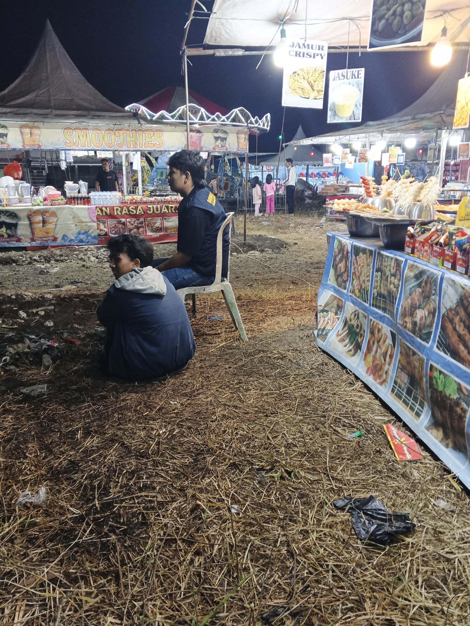 This image captures a nighttime scene at a food stall area, likely a night market, in Sukabumi, Indonesia. In the foreground, two young males are seated on the ground. One is facing away from the camera, his back hunched, wearing a dark blue hooded jacket with gray trim. The other, seated on a plastic chair, is wearing a dark blue shirt with yellow accents and jeans, facing the food stalls.
Behind them, several food stalls are visible. One stall displays a banner that reads "SMOOTHIES" with images of fruit smoothies. Another stall has a banner with text that includes "AN RASA JUAR" and images of various food items such as skewers, kebab, and what appears to be fried mushrooms. Signs overhead advertise "JAMUR CRISPY" and "JASUKE".
The environment consists of dry, scattered grass and some debris on the ground. The stalls are covered by tents, and lights illuminate the area. In the background, a few other people are visible near the stalls, including a man and two young girls. The overall atmosphere suggests a casual, outdoor food gathering place.