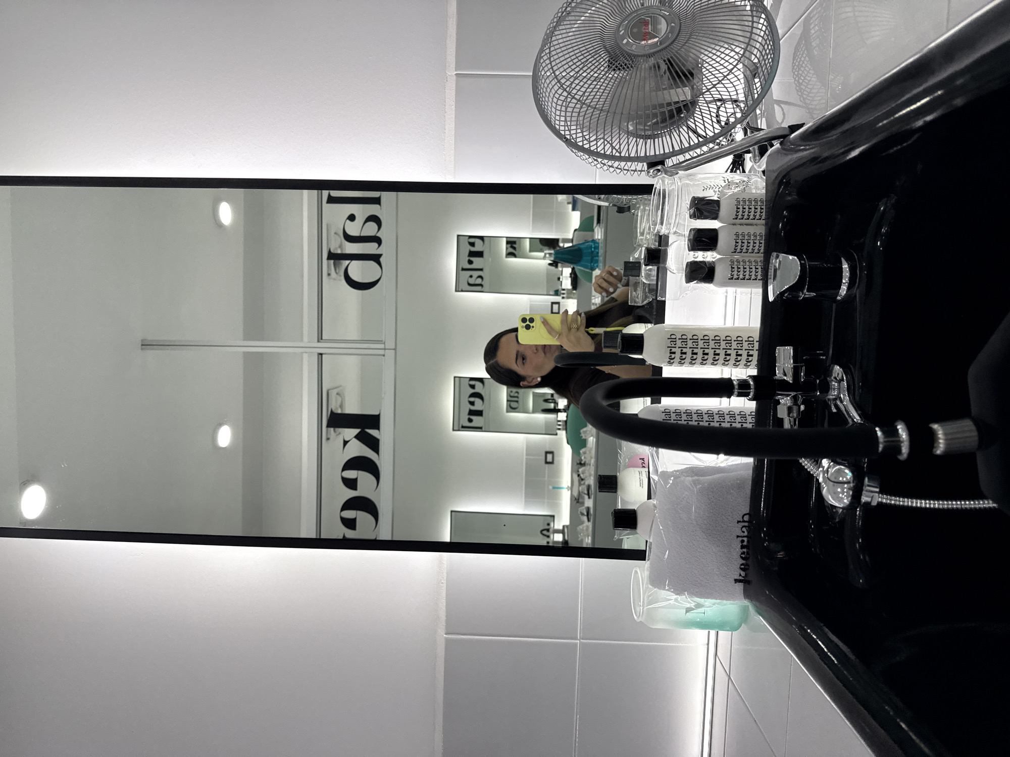 Here is a forensic description of the image.
The image shows a person reflected in a mirror in a hair salon. The salon's logo, "KGG dsl," is visible on the mirror. The person is holding a yellow phone, presumably taking a selfie. In the foreground, there is a black hair washing station with a white towel that says "keerlab" on it. There is a standing fan to the side of the washing station and various bottles of "keerlab" products behind the washing basin. The environment is clean and brightly lit, with white tiled walls.