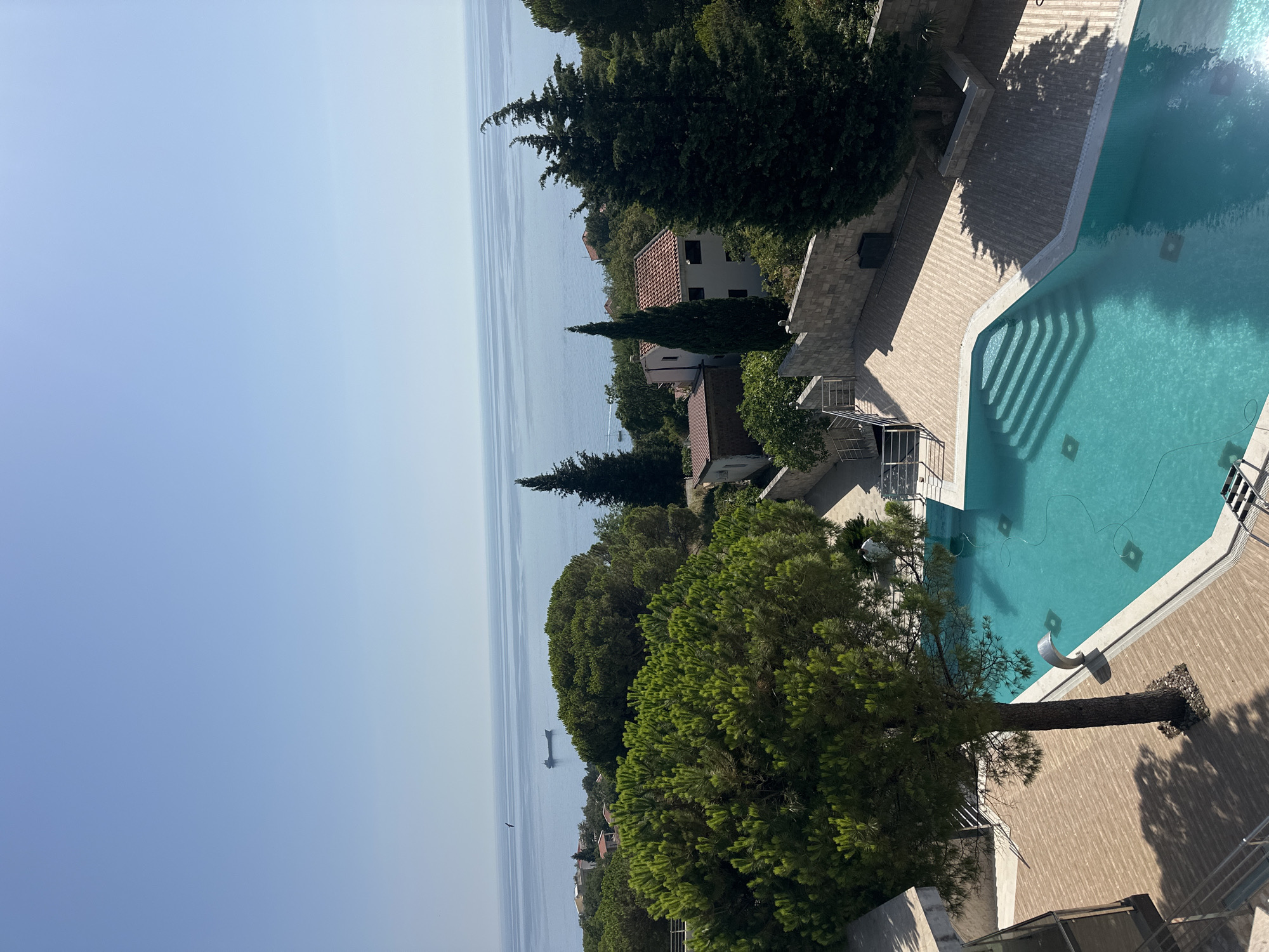This image captures a serene outdoor scene in Brca, Montenegro. In the foreground, a bright turquoise swimming pool with a stepped entrance occupies the lower right portion of the frame. The pool is surrounded by a light-colored tiled deck. To the left of the pool, a mature tree with dense green foliage stands on the deck, casting a shadow.
Beyond the pool and deck, lush green trees and vegetation dominate the mid-ground. Several buildings with terracotta roofs are visible nestled amongst the trees. In the distance, the calm, blue Adriatic Sea stretches towards the horizon under a clear, pale blue sky. A cargo ship and a small bird are visible on the water. The overall impression is one of tranquility and natural beauty.