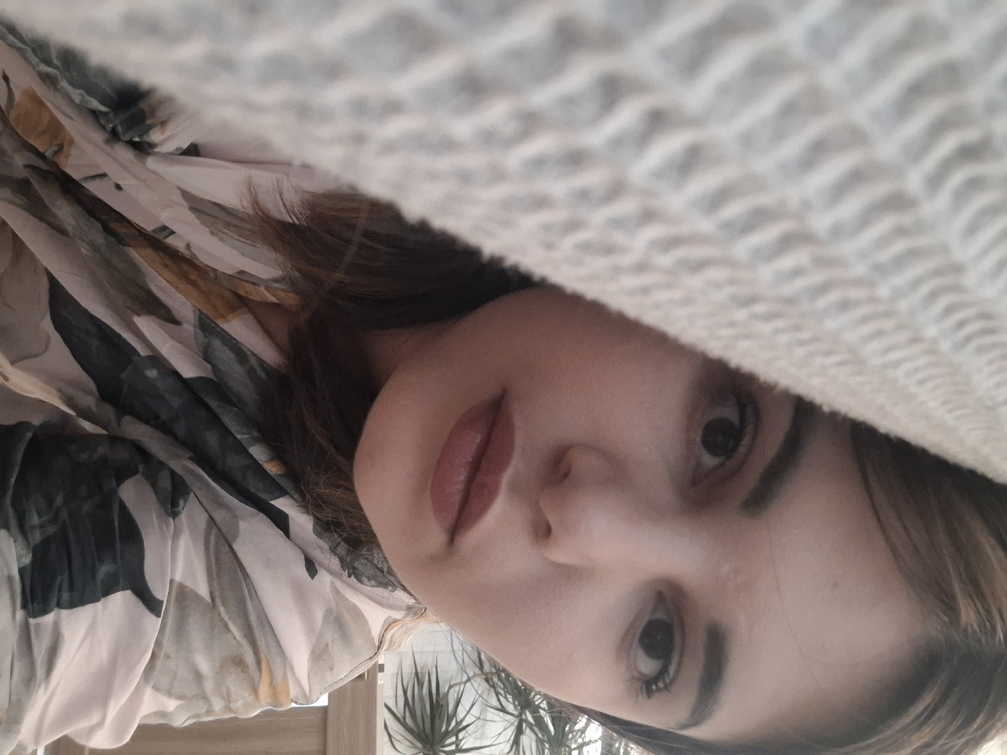 A woman's face is the focal point, partially obscured by a white, textured blanket above. The woman's complexion is fair, with visible eyelashes and eyebrows. Her lips are a muted pink. She is wearing a patterned garment. Behind her, partially visible in the background, is greenery. The location is unidentifiable from the given image.