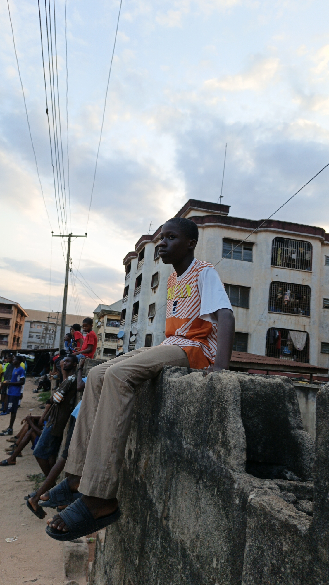 This image captures a candid street scene in Onitsha, Nigeria, during late afternoon or dusk, as indicated by the soft, warm light and the sky transitioning from light blue to orange-pink with scattered clouds.
The central subject is a young Black boy, likely a teenager, seated on a rugged, grey concrete wall in the foreground. He is positioned facing right, with a thoughtful or contemplative expression as he looks out of the frame. He wears an orange and white striped short-sleeved t-shirt with a faint, unclear yellow logo or text on the front, light brown or khaki trousers, and dark blue slide sandals on his feet, which dangle off the wall.
To the left and slightly behind him, several other people, predominantly young males, are visible. Some are sitting on the ground or low structures, while others are standing, suggesting a casual gathering or simply people socializing in the area.
The background reveals a typical urban environment. A prominent utility pole with numerous overhead power lines stretches across the midground on the left. Behind the boy, a multi-story, light-colored building with signs of wear stands out, featuring multiple windows, some barred, and visible items like laundry on balconies. Other multi-story buildings are discernible further in the distance. The ground appears to be unpaved or dirt in sections.
The scene provides a snapshot of daily life in a Nigerian city, characterized by its unique architecture, infrastructure, and the informal gathering of people. No clearly legible text is visible other than the faint logo on the boy's shirt.