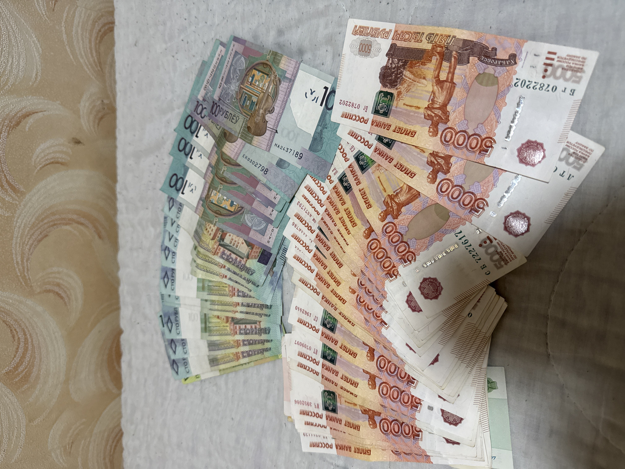 The image shows two piles of Russian currency lying on a surface. On the left is a stack of 100 Ruble banknotes. On the right is a larger stack of 5000 Ruble banknotes. The scene appears to be staged, showcasing the money. The presence of the Russian Ruble suggests a location or association with Russia, but it doesn't confirm a location. There's no confirmation of the city of Лабжанскі сельскі Савет, Belarus.