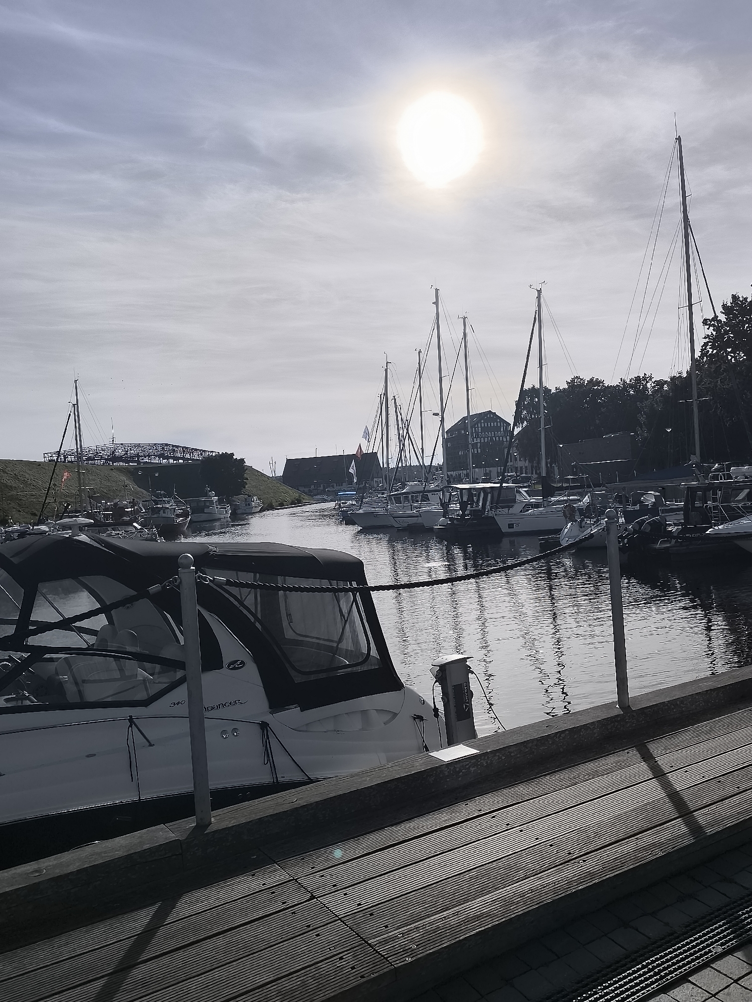 The image depicts a harbor scene in Klaipėda, Lithuania, likely at the Old Town harbor or a nearby marina, on a bright, partly cloudy day. In the foreground, the edge of a wooden boardwalk leads the eye towards the water, where several boats are docked. The nearest vessel is a white motorboat with a black canopy and clear windows, moored alongside the boardwalk. It has a railing and rope details visible. Beyond this boat, the water is calm and reflects the sky and the masts of sailboats.
A line of sailboats with tall masts stands moored further out in the harbor. These sailboats have white hulls and rigging, and some have flags visible. In the middle ground, several buildings with traditional European architecture, including a building with a darker facade and red-tiled roof, are visible along the waterfront. Behind them, on a raised grassy embankment, a complex structure resembling a bridge or elevated walkway can be seen. The sky is a pale blue with wispy white clouds, and a bright sun is prominently positioned in the upper left quadrant of the image, casting a slight glare and creating soft shadows. The overall impression is one of a peaceful, well-maintained harbor on a sunny day. There are no people clearly visible in the image.