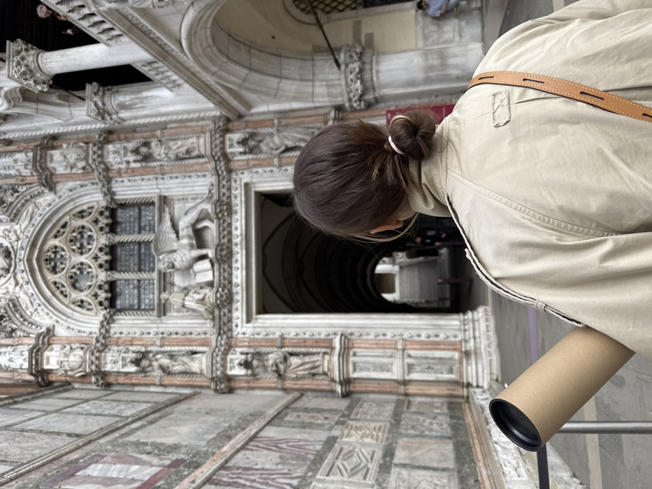 An individual is depicted from behind, centered in the lower frame, observing the Porta della Carta, the ceremonial entrance to the Doge's Palace (Palazzo Ducale), in Venice, Italy. The person has brown hair secured in a low bun with a light pink hair tie and wears a light tan collared jacket or trench coat. They carry a cylindrical brown cardboard tube, capped in black, under their left arm. A light brown leather strap is visible over their right shoulder.
The Porta della Carta, constructed from white, pink, and gray marble, dominates the background. It features a grand Gothic archway adorned with intricate carvings, including a rose window, multiple statues in niches along its vertical piers, and a prominent relief sculpture above the entrance depicting the winged Lion of St. Mark flanked by figures (historically the kneeling Doge Francesco Foscari and another figure). The archway opens into a deep, vaulted passage with stairs visible ascending in the distance. To the left of the gate, a stone wall exhibits horizontal bands of varied marble and geometric relief patterns. To the right, sections of the palace's arcaded architecture, including columns and an upper-level balustraded balcony, are visible. Two indistinct figures are discernible within the wider palace structure: one on the upper balcony and another seated further back inside the arched walkway.