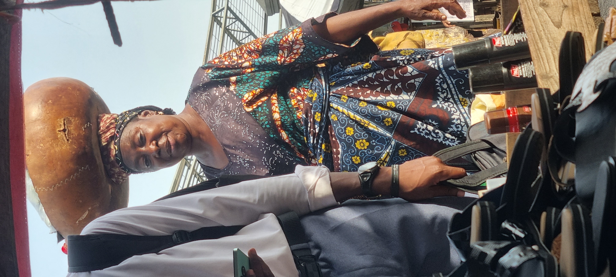 A middle-aged woman with a large gourd on her head looks down at a customer, possibly in a marketplace in Lagos, Nigeria. The woman wears a patterned dress and headwrap. The customer, wearing a white shirt, holds a cell phone in their hand, their arm visible at the bottom of the frame. The scene is cluttered with merchandise, including black sandals, shoe polish, and wooden supports. The background hints at an urban environment, possibly a busy street or market.