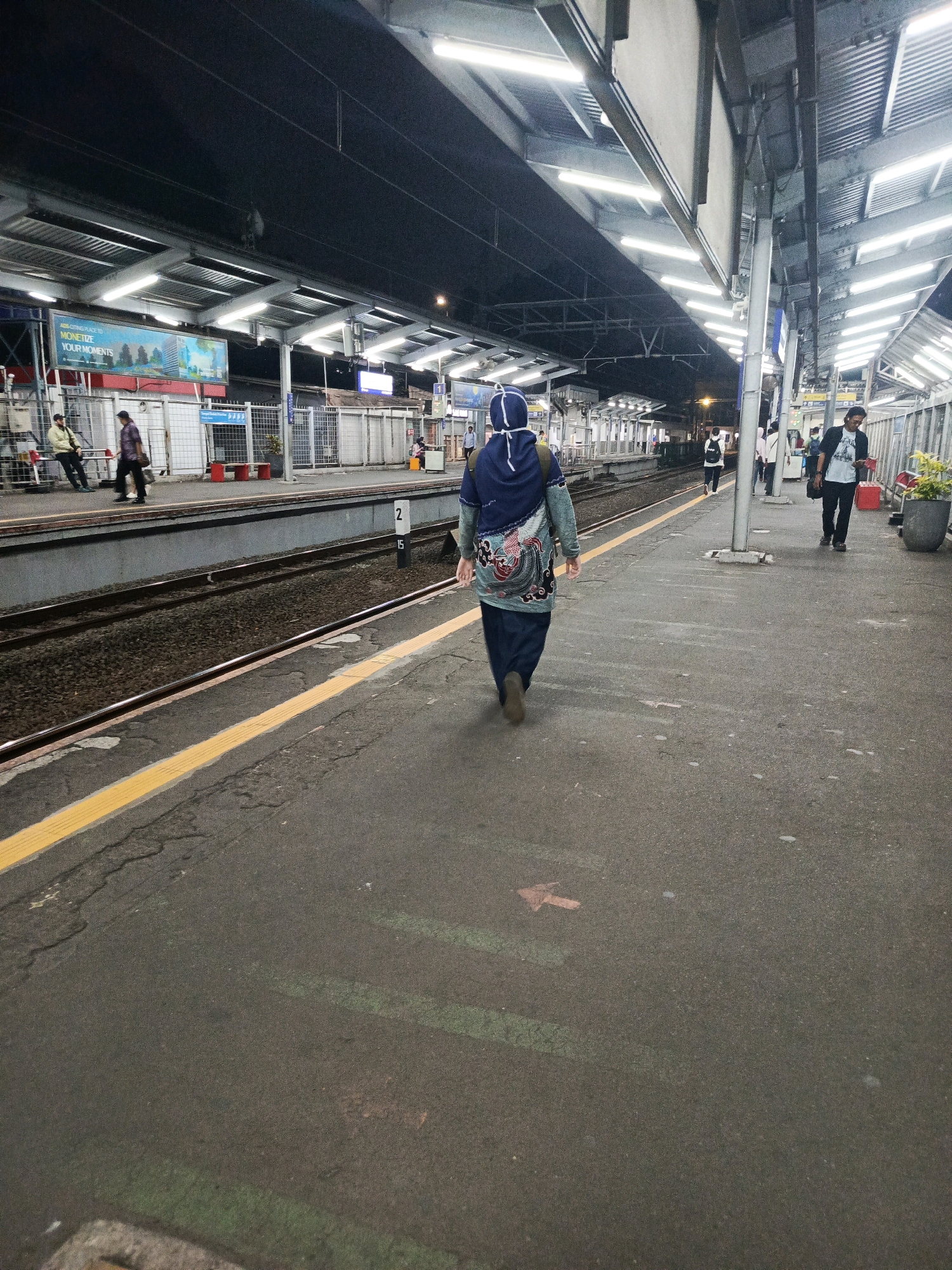 The image shows a train station platform at night in South Jakarta, Indonesia. A person wearing a blue hijab and a patterned jacket is walking away from the camera on the platform. The platform is paved and has yellow tactile paving along the edge. In the background, there are train tracks, other people walking, and a covered waiting area with lights. A sign with the number "2" is visible near the tracks. The environment appears to be outdoors, under an illuminated canopy structure.