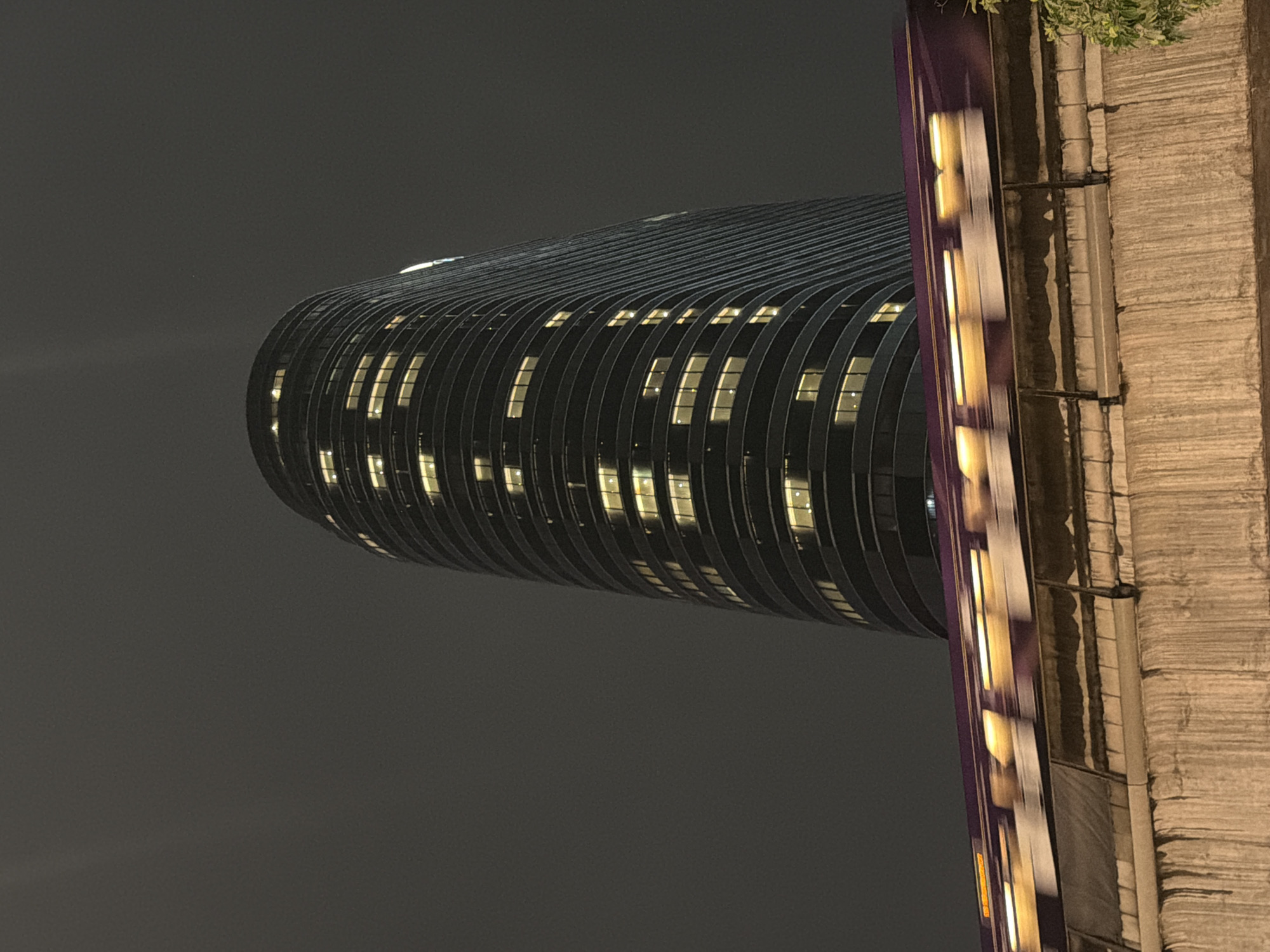 An upward-angle, night-time photograph captures a blurry, purple train moving left to right across the frame, its windows emitting streaks of yellow light. The train passes along an elevated track supported by a textured, concrete structure in the foreground. Behind the track, a tall, cylindrical skyscraper with many illuminated windows rises against a dark, cloudy sky. The building's facade appears to be made of dark material with horizontal banding. The overall scene suggests an urban environment in Bangkok, Thailand, at night.
