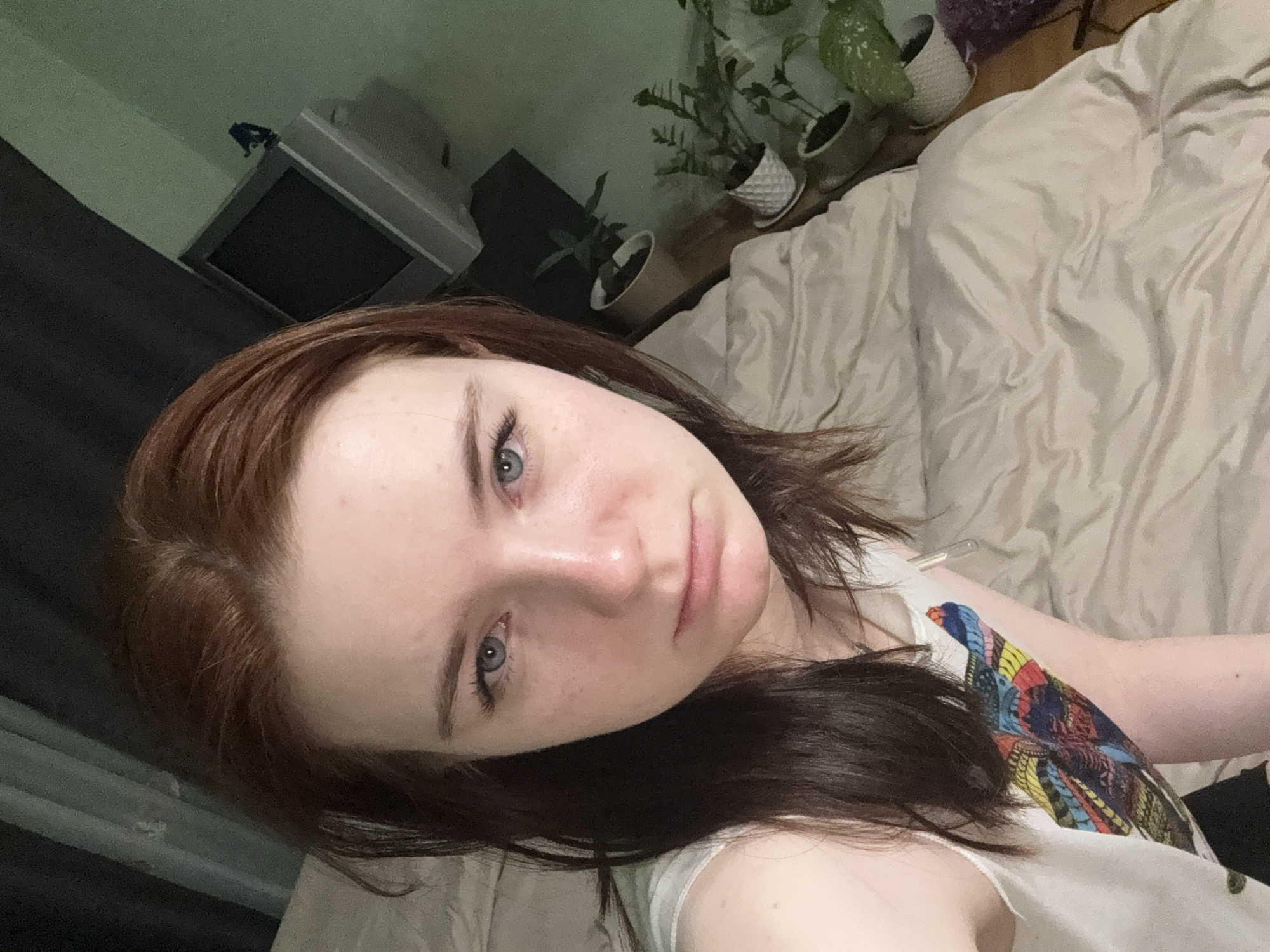 A close-up, tilted shot features a young woman with light skin, blue eyes, and brown hair with red highlights. She is wearing a white shirt with a colorful design. Behind her is a bed with light-colored bedding. In the background, several potted plants are visible on a surface near a television. The location is Unknown, Unknown.