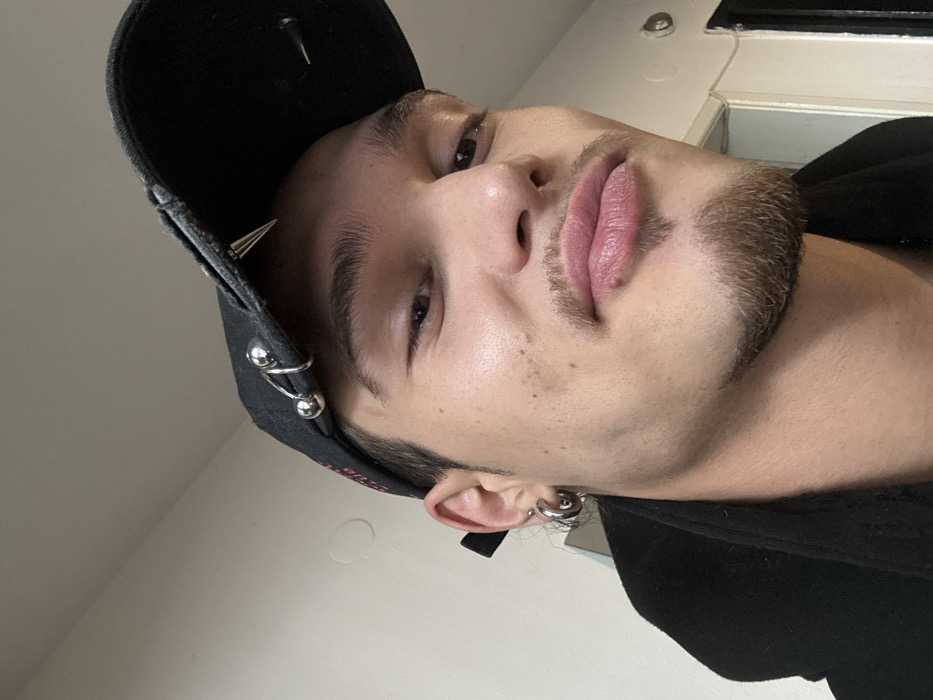 A young male individual is depicted in a close-up, chest-up indoor portrait. He has light skin, dark eyes, and dark eyebrows. A light mustache, soul patch, and chin strip are present. His lips are full and slightly pursed. A small mole is visible on his lower lip. He wears a black baseball cap backward, adorned with two silver-colored spikes and two silver-colored rings on what would be the front brim. A silver-colored hoop earring is visible in his left earlobe. He is wearing a dark-colored garment, possibly a jacket or hoodie. The individual looks directly towards the viewer. The background consists of a plain, light-colored wall. To the right, a dark door frame or opening is partially visible. No specific architectural or environmental features are present to identify the precise location within Sofia, Bulgaria, beyond an indoor setting.