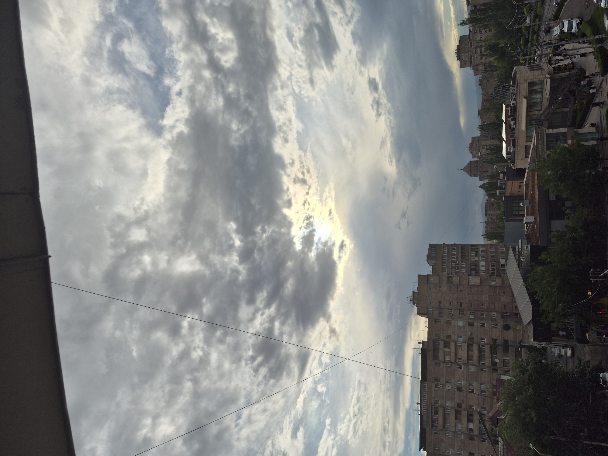 The image presents an urban landscape under a dramatic, cloudy sky, captured from an elevated viewpoint, likely a balcony or a high-rise window. A dark, solid structure forms the left edge of the foreground.
The sky is a dominant feature, filled with a diverse array of clouds, ranging from dense, dark grey formations to lighter, wispy white streaks. Patches of lighter blue sky are visible between the cloud masses. The sun, partially obscured by clouds, creates a bright, almost golden glow as it breaks through a gap in the upper center of the frame, suggesting a lower angle, possibly late afternoon or early evening. Several thin utility wires crisscross the sky, adding an element of urban infrastructure.
Below the sky, the cityscape unfolds with numerous multi-story buildings. These structures, characterized by their concrete facades and repetitive window arrangements, bear the hallmarks of post-Soviet architecture common in cities like Yerevan, Armenia. Trees and green foliage are scattered throughout the urban environment, providing natural accents amidst the concrete. In the middle and far distance, more buildings line streets where parked and moving vehicles are visible, along with a few indistinct human figures. A notable architectural detail in the distant background is a large building with a distinctive pointed roof or spire, possibly a church or cathedral, adding a landmark to the horizon.
The overall scene depicts a typical urban environment with no specific event or intense activity taking place. The time of day is clearly daylight, indicated by the visible sun, and the cloud cover suggests a dynamic weather pattern. No clearly readable text is discernible within the image.