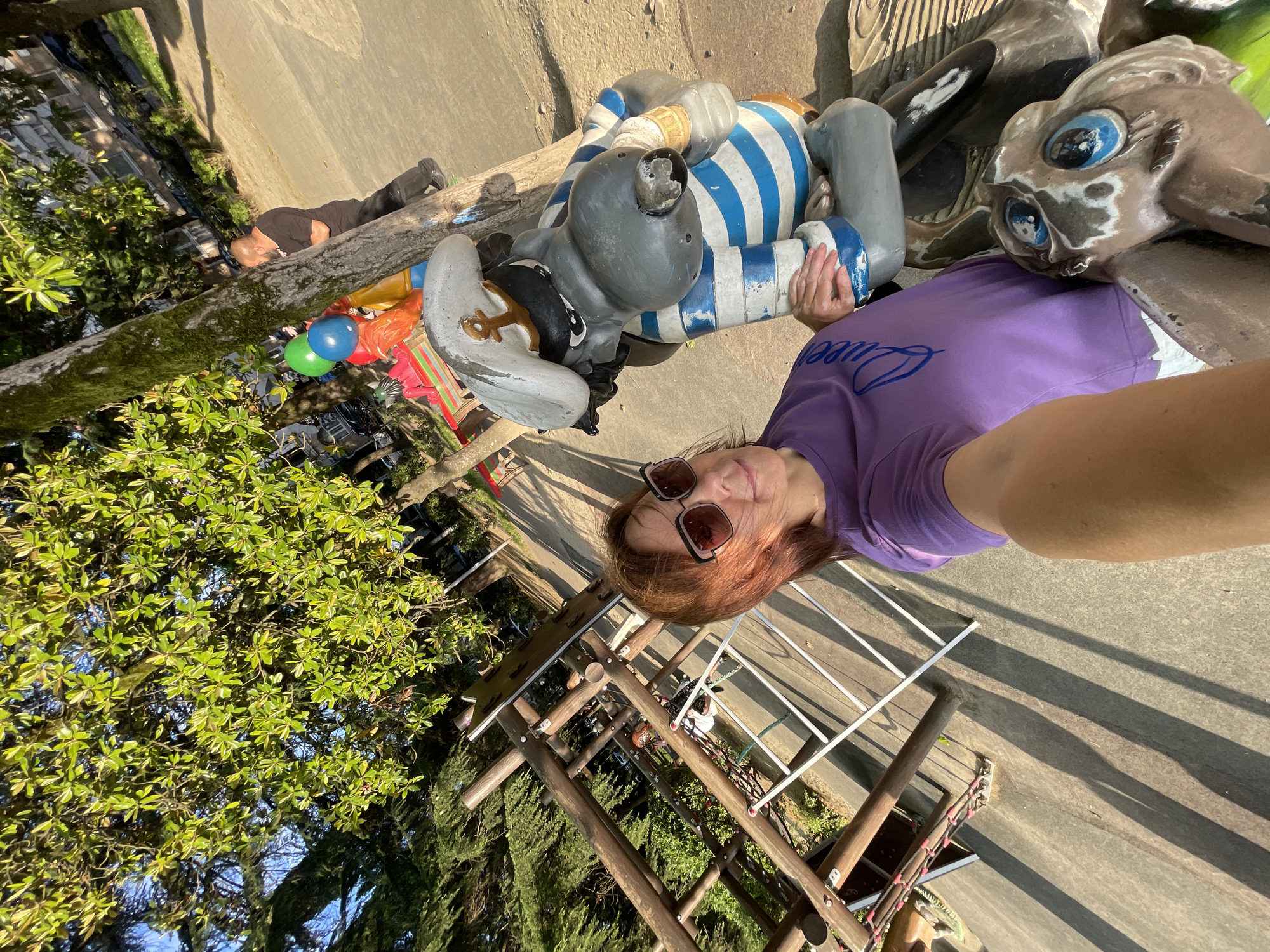 A woman in a purple t-shirt and sunglasses takes a selfie with a statue of a cartoon character in a sailor's hat and striped shirt. The statue appears to be part of a playground or park attraction, with a wooden climbing structure visible in the background. The location is specified as Sochi, Russia. The scene is outdoors, with trees and a pathway. The lighting suggests it is daytime, possibly late afternoon due to the long shadows.