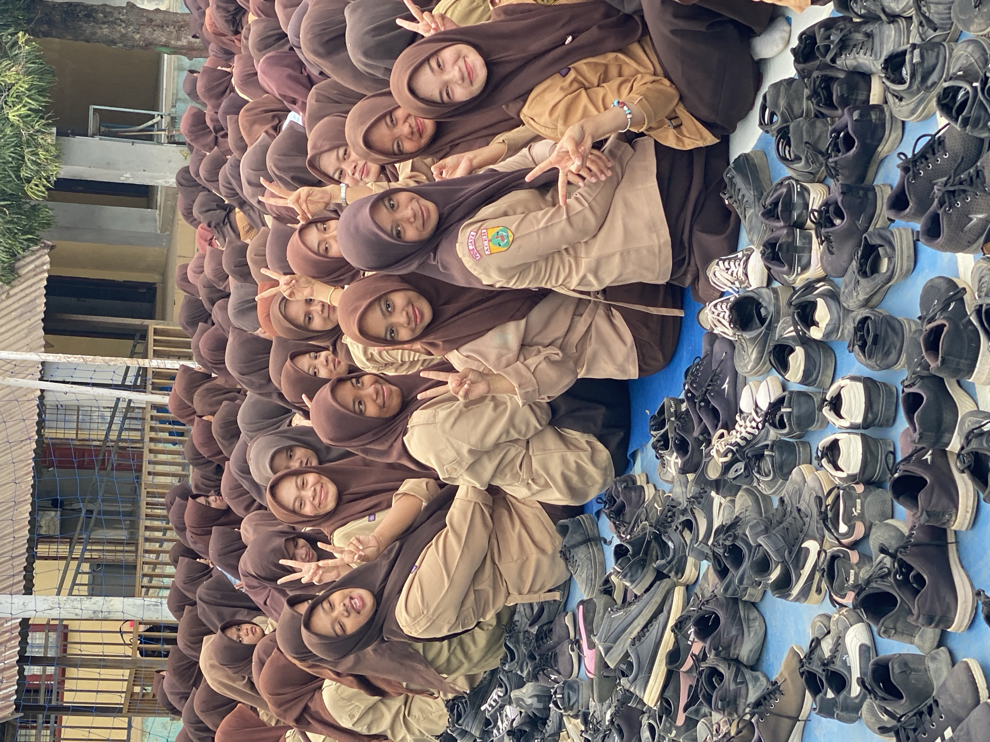 A group of young women, likely students, are gathered outdoors. They are wearing brown hijabs and tan-colored uniforms, suggesting a school setting. Many of them are smiling and posing for the camera, with some making peace signs with their fingers. They are sitting on a blue tarp, which is covered with a large collection of black and white sneakers. The sneakers are neatly arranged in rows, indicating that they may have been removed for an activity or event. In the background, there are buildings with corrugated metal roofs and a fence, further supporting the idea of an outdoor school or communal area. The overall atmosphere appears cheerful and communal.