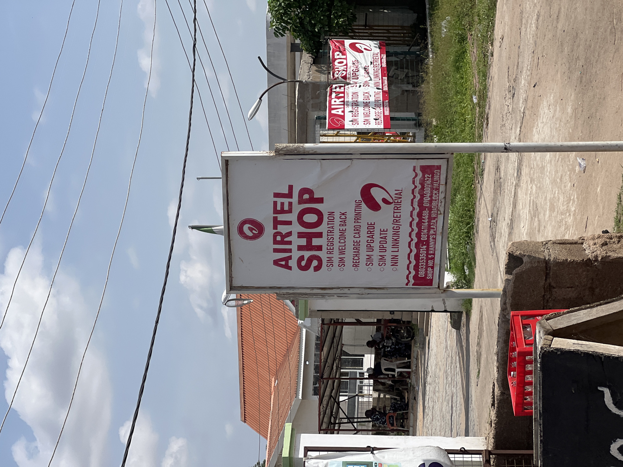 A daytime, eye-level, medium shot captures an Airtel shop storefront in Jalingo, Nigeria. The main focus is a large, white sign with red lettering that reads "AIRTEL SHOP" prominently displayed. Below the main title, a list of services is provided: "SIM REGISTRATION," "SIM WELCOME BACK," "RECHARGE CARD PRINTING," "SIM UPGRADE," "SIM UPDATE," and "NIN LINKING/RETRIEVAL." Contact information and shop number are also visible at the bottom of the sign.
To the right of the main sign, a smaller, red Airtel sign is visible, featuring similar services. A street lamp stands between the two signs. In the background, buildings with orange tiled roofs are visible, suggesting a commercial or residential area. Several people are seated under a shaded structure behind the Airtel shop, some appearing to be customers or staff. A motorcycle is parked nearby.
The environment appears to be an outdoor setting with a paved ground surface. A red crate of empty bottles sits in the foreground, to the left of the main sign. The sky is partly cloudy, indicating daylight. The overall scene depicts a typical retail establishment offering mobile services in a Nigerian city.
