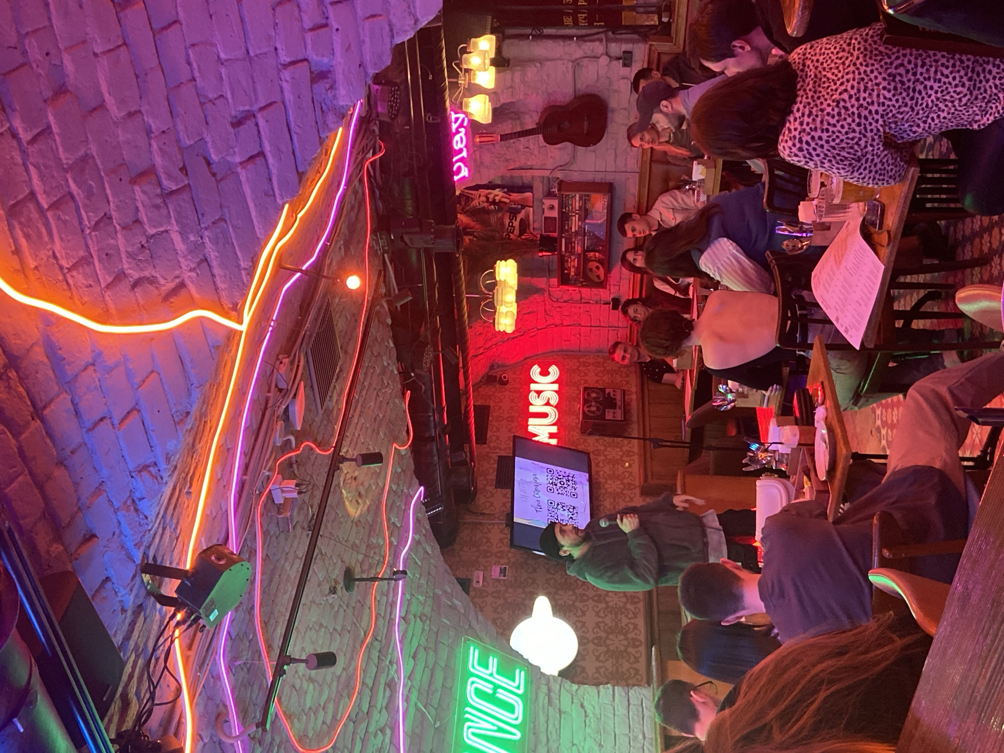 This media file captures a vibrant, dimly lit indoor venue in Saratov, Russia, likely a bar or club, during an evening event. The scene is dominated by numerous young adults gathered around tables, enjoying the atmosphere. The image is filled with the warm glow of various neon lights in orange, pink, red, and green, illuminating exposed brick walls and an arched ceiling.
In the foreground and midground, people are seated at dark wooden tables with drinks and menus. Many are looking towards the front of the room, where a person stands holding a microphone, suggesting an ongoing performance or a hosting role. A flat-screen monitor mounted on the wall displays QR codes and Cyrillic text, which upon closer inspection, appears to read "Твои секреты?" (Your secrets?) and "Свободный микрофон" (Open mic), strongly indicating an open mic night, karaoke session, or interactive event.
Notable decorative elements include an acoustic guitar mounted on a brick wall, various vintage-style light fixtures, and distinct neon signs spelling out "PLAY" in pink, "MUSIC" in red, and "DANCE" in green. The overall ambiance is lively and social, with individuals engaging in conversation, watching the stage, and enjoying beverages. The time of day is clearly evening or night, relying entirely on artificial, atmospheric lighting. The venue exhibits an eclectic decor with a mix of brickwork, wooden furniture, and modern neon art.