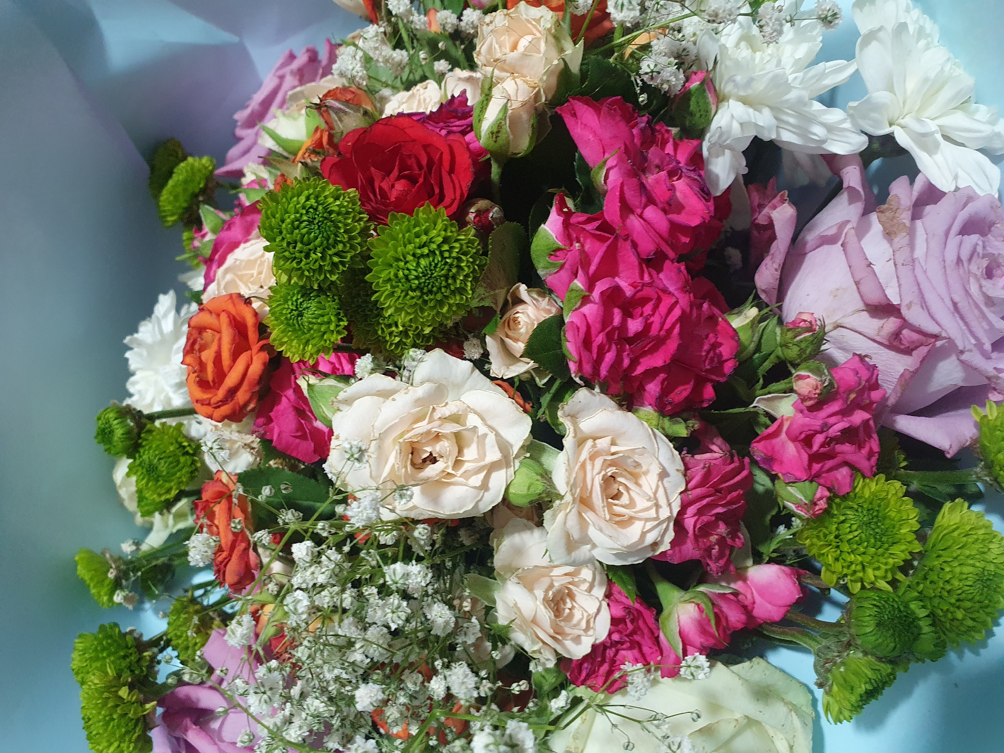 A bouquet of flowers is arranged in a close-up shot against a light blue backdrop. The bouquet features a variety of flowers including pink roses, white roses, orange roses, red roses, green chrysanthemums, and small white filler flowers. The lighting casts shadows, highlighting the textures and layers of the petals.