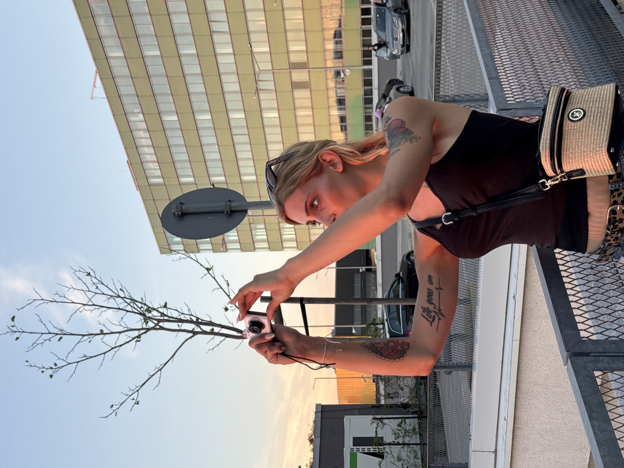 In an outdoor urban setting, a young woman with blonde hair and sunglasses perched on her head is captured from the chest up, holding a small digital camera to take a photo. She is wearing a dark tank top and has tattoos on her arms, including one on her left arm that reads "Life goes on" with a heartbeat graphic. A straw crossbody bag with a black strap is slung over her shoulder.
The scene appears to be during the golden hour, with warm, soft light illuminating the woman's face and the background. The setting includes a large, modern building with a facade of horizontal windows. In the background, cars are visible on a street, suggesting an urban environment. A bare tree branch with a few small leaves extends into the frame from the left, reaching towards the sky. The overall mood seems casual and relaxed, with the woman focused on capturing a moment with her camera. The image is taken in Sofia, Bulgaria.