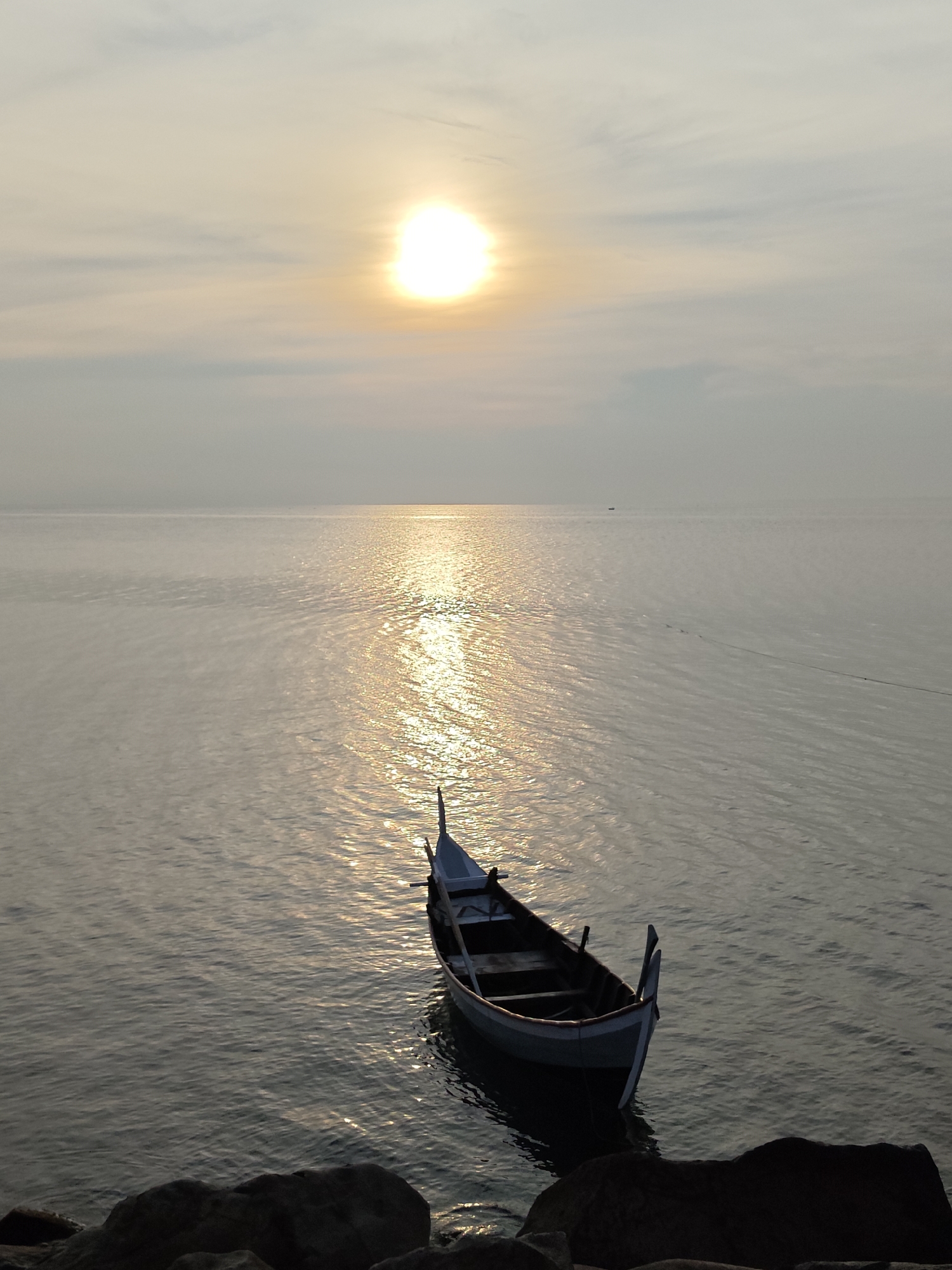 This image captures a serene coastal scene in Lhokseumawe, Indonesia, at either sunrise or sunset. A single, traditional-style wooden boat, painted in light blue/grey and dark brown, floats gently on calm, reflective water, positioned towards the lower right of the frame.
The bright, intense sun is a prominent feature in the upper-middle sky, casting a dazzling path of shimmering golden light across the water, extending directly towards the viewer. The sky is soft and hazy, with delicate, wispy clouds that diffuse the sunlight, creating a tranquil atmosphere. Dark, indistinct rocks or landmass are visible in the foreground, suggesting the boat is moored near the shore.
In the far distance, a tiny, faint silhouette of another boat can be seen on the horizon, underscoring the expanse of the water. A thin, dark line, possibly a rope or fishing line, stretches across the mid-ground of the water. The scene evokes a sense of peaceful solitude and the quiet beauty of nature. There is no visible text within the image.