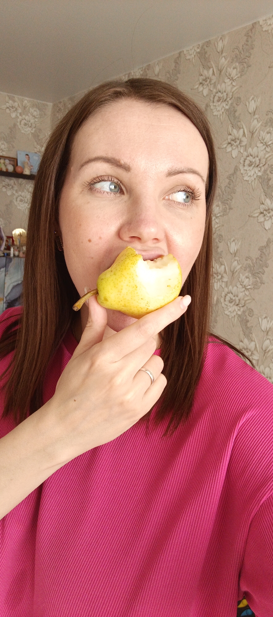 A woman is depicted in a close-up, holding a yellow pear with a bite taken out of it near her mouth. She has long brown hair, parted centrally, and light blue-green eyes that are looking upwards and to her right, away from the camera. Her complexion is light, and she appears to have natural makeup, with visible eyelashes. She is wearing a vibrant, fuchsia-pink top with a distinct ribbed texture. A simple silver band ring is visible on the ring finger of her left hand, which is gently supporting the pear.
The setting is indoors, with a light-colored wall featuring an ornate, slightly raised floral or leafy pattern wallpaper visible in the background. A small portion of a light-colored ceiling is also visible towards the top. On the left side of the frame, a hint of a shelf with a few indistinct decorative items, including what appears to be a small framed picture, can be seen.
The lighting is bright and natural, suggesting it is daytime. Her expression is contemplative or observant, given her gaze. The location context is Chaikovsky, Russia. There is no visible text in the image.