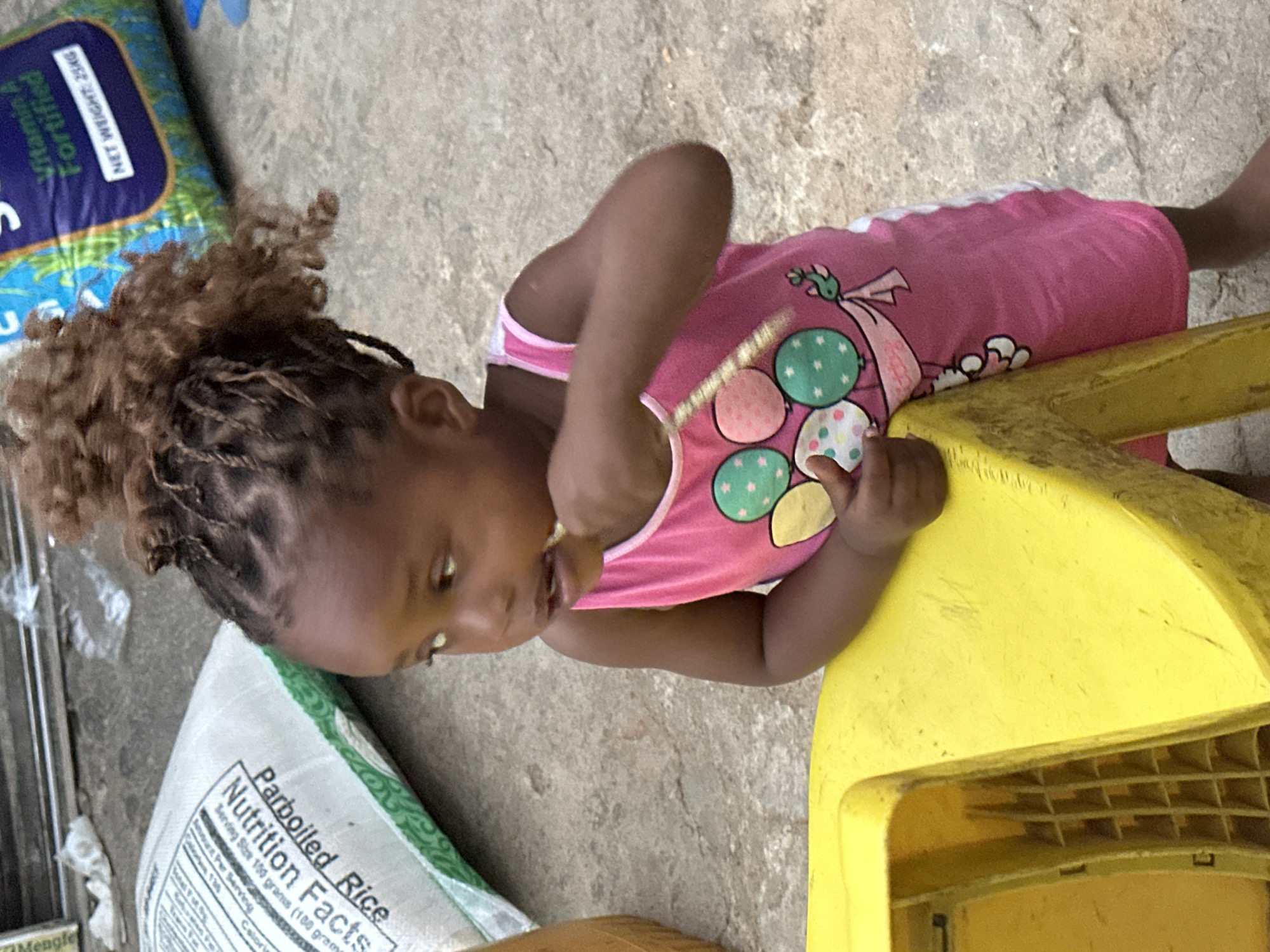 A young, dark-skinned child, possibly a girl, with braided hair and curly sections on top, is captured in a close-up, high-angle shot outdoors in Narayi, Nigeria. She is wearing a pink tank top with white trim, featuring a colorful design of balloons and a cartoon character flying. The child is positioned next to a bright yellow plastic object, which appears to be a child's chair or toy, and is engaged in an activity where she brings her right hand to her mouth, suggesting she is eating or playing.
The scene unfolds on a concrete surface under natural daylight. In the background, two large bags are visible. One blue and green bag has text indicating "Vitamin A Fortified" and "NET WEIGHT: 25KG", with "Mengle" also partially visible. Another white and green bag prominently displays "Parboiled Rice" and "Nutrition Facts", with "Serving Size 100 grams (100 gram)" also decipherable. The child's expression is one of focus on her immediate activity.