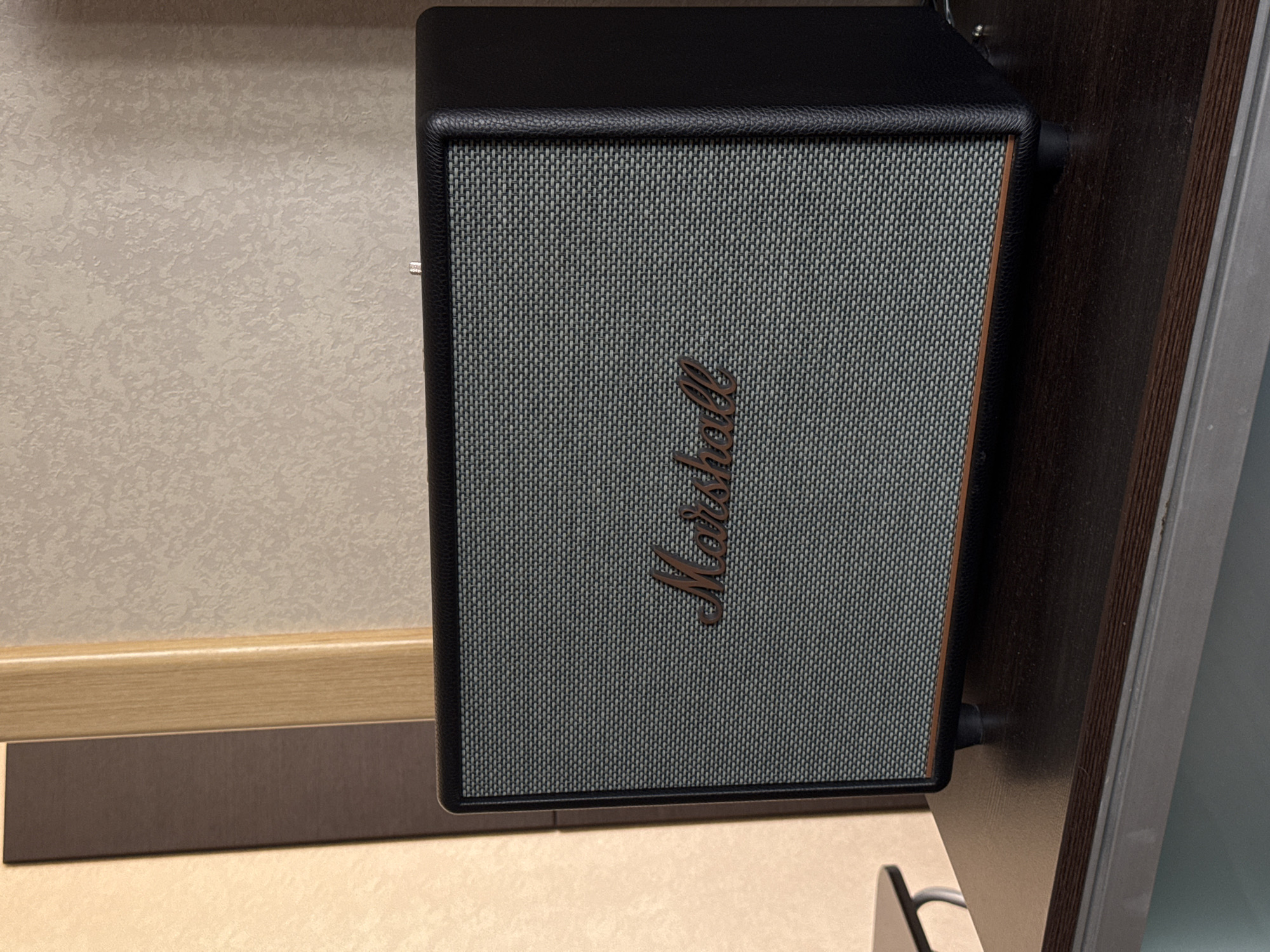 A Marshall speaker with a black exterior and a grey mesh grill is placed on a dark wood surface. The Marshall logo is visible in a bronze color on the grill. The background consists of a textured wall and a wooden door frame. The scene is indoors and appears to be a domestic or office setting. There are no people or animals present in the image. The lighting is soft, suggesting ambient indoor lighting. There is no indication of the specific location being Yekaterinburg, Russia.